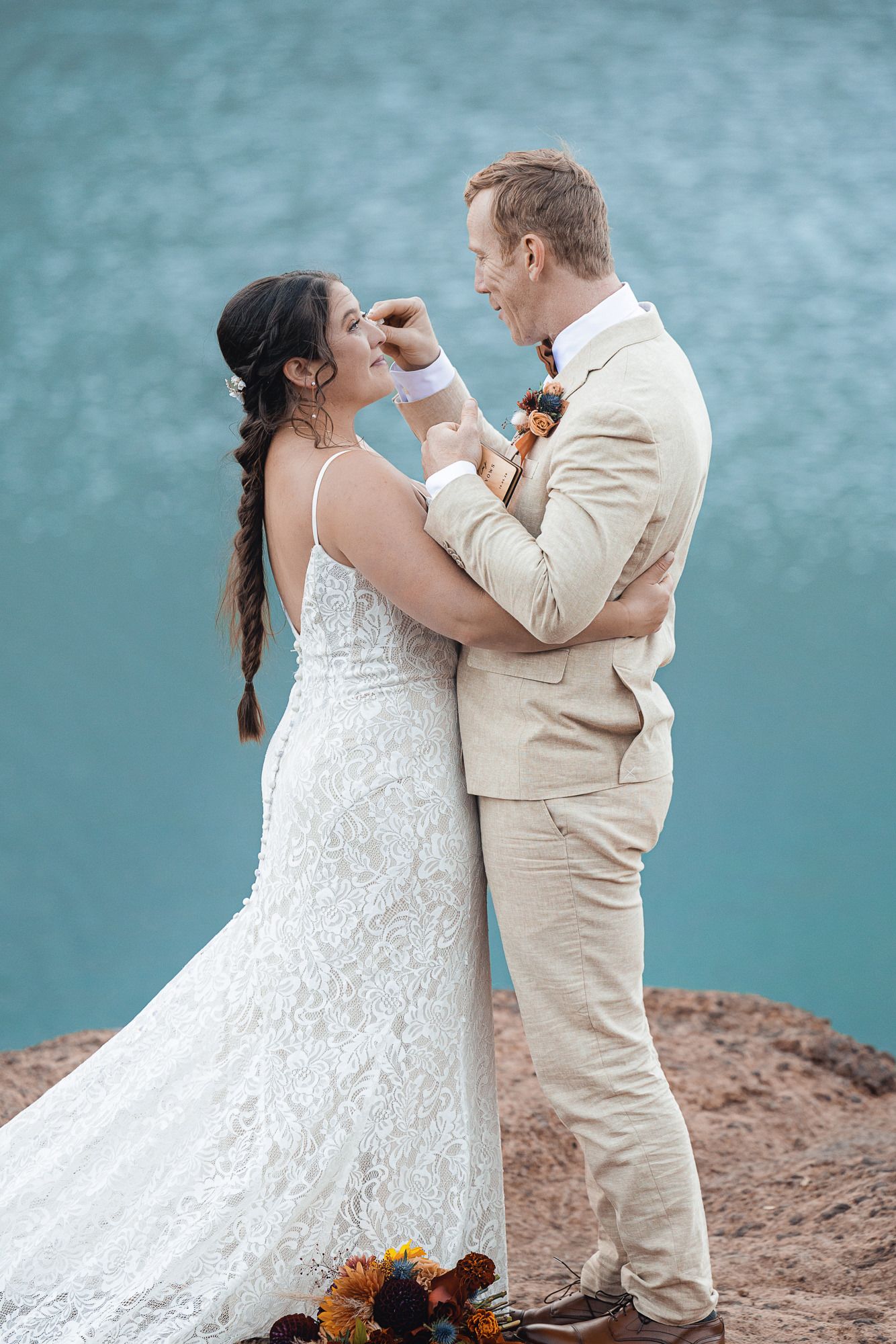 Bride in a lace wedding gown with a long braid embraces a groom in a tan suit on a rocky cliff above turquoise water.