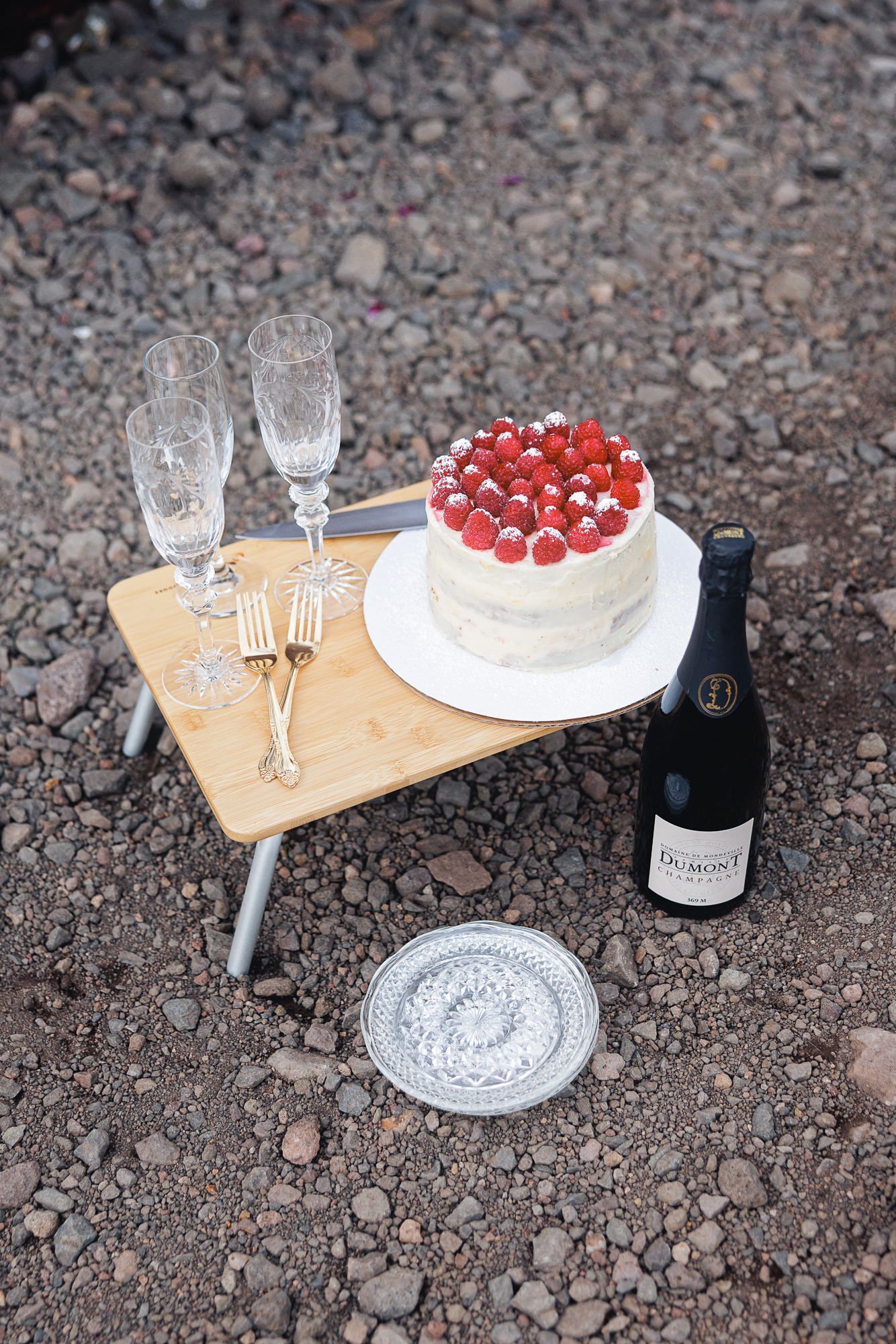 Raspberry-topped cake, champagne bottle, crystal flutes and gold forks on a small wooden picnic table set on volcanic gravel.