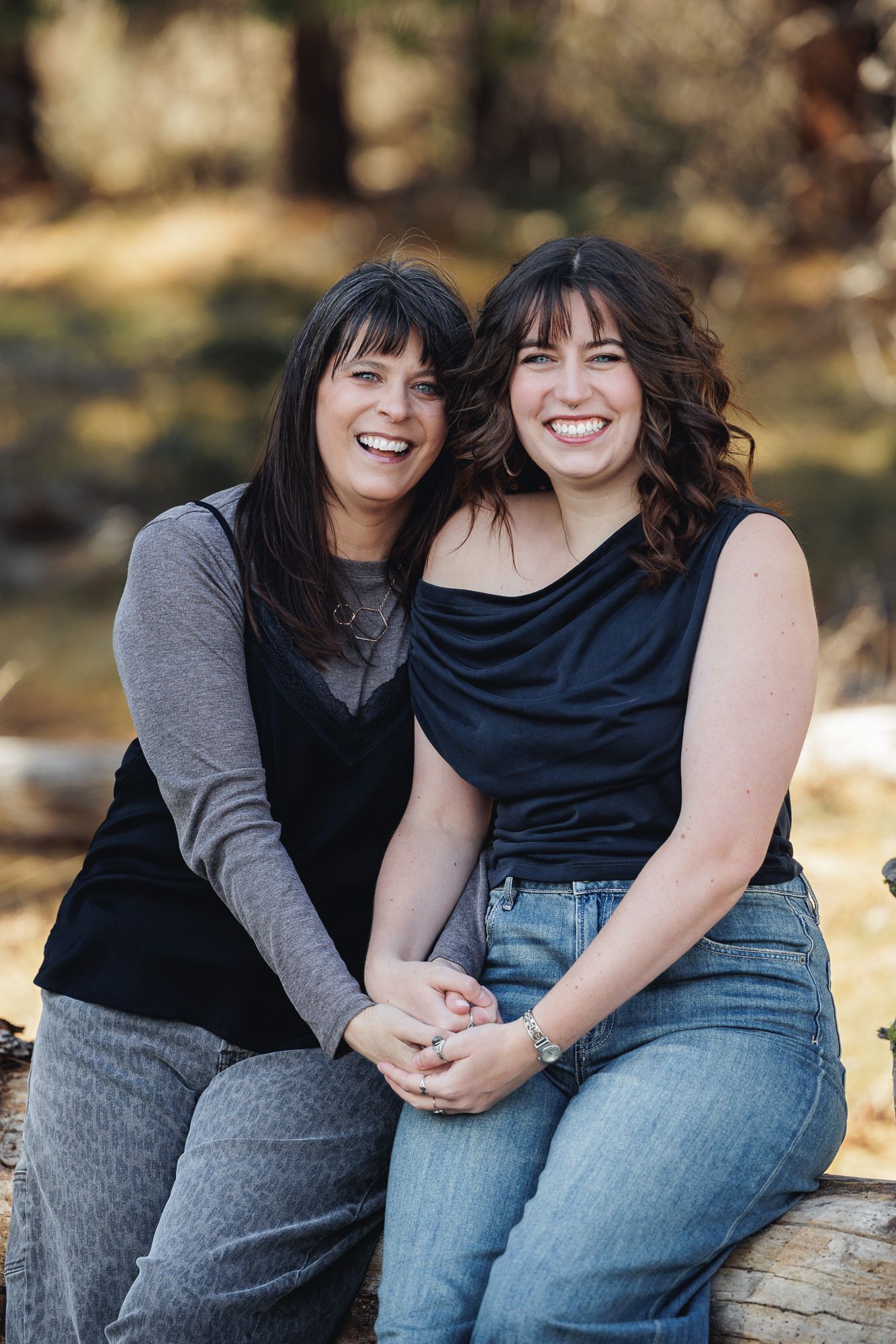 Two smiling women with dark hair sit together outdoors on a log, wearing casual clothing in a wooded setting.