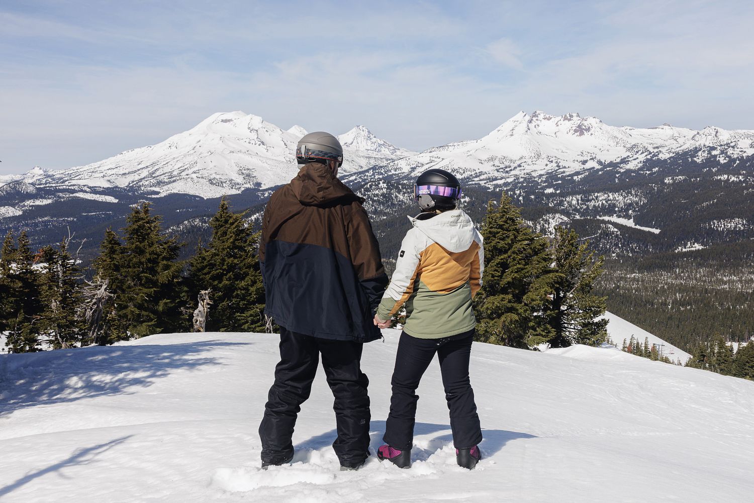 Two people stand on a snowy mountain slope with snow-capped peaks in the background.