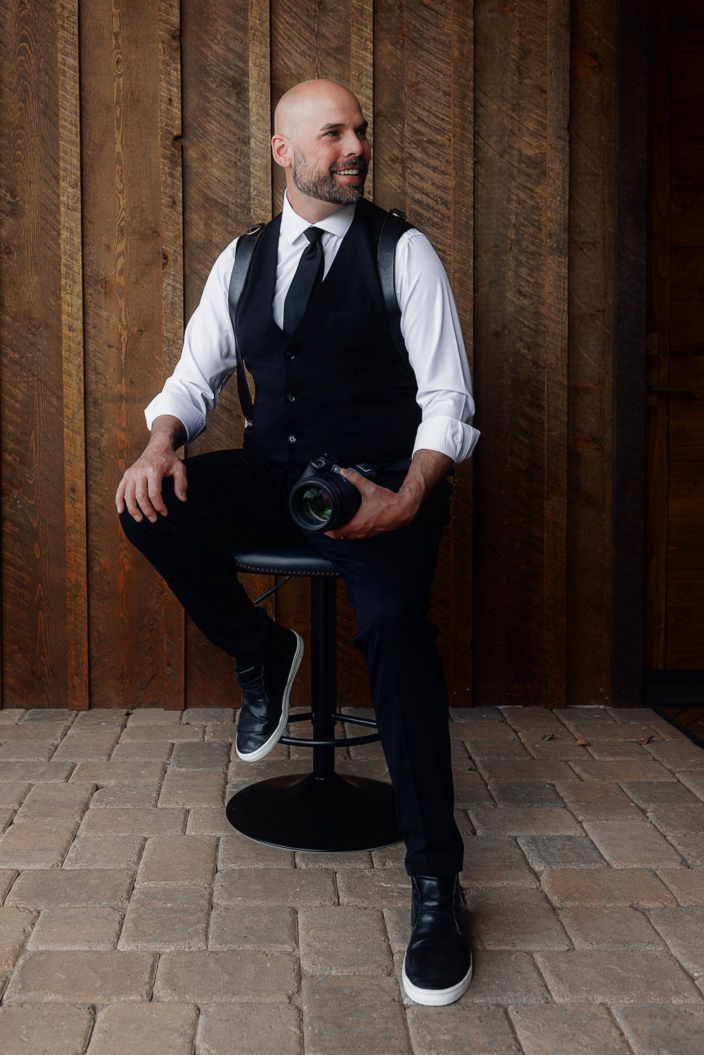 A person in formal attire sits on a chair against a rustic wooden wall and brick floor.