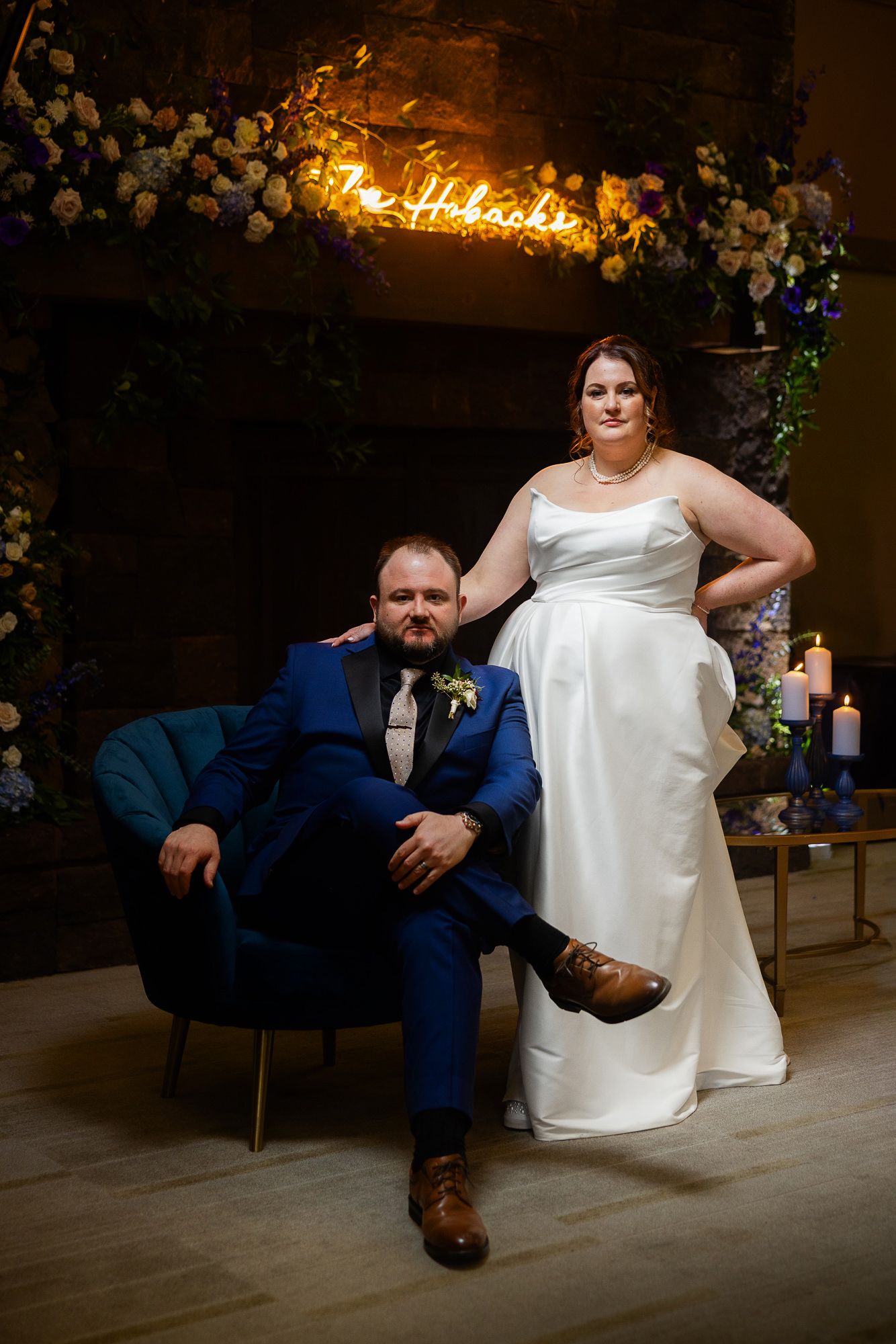 A couple in wedding attire relaxes on a blue velvet chair during their reception celebration.