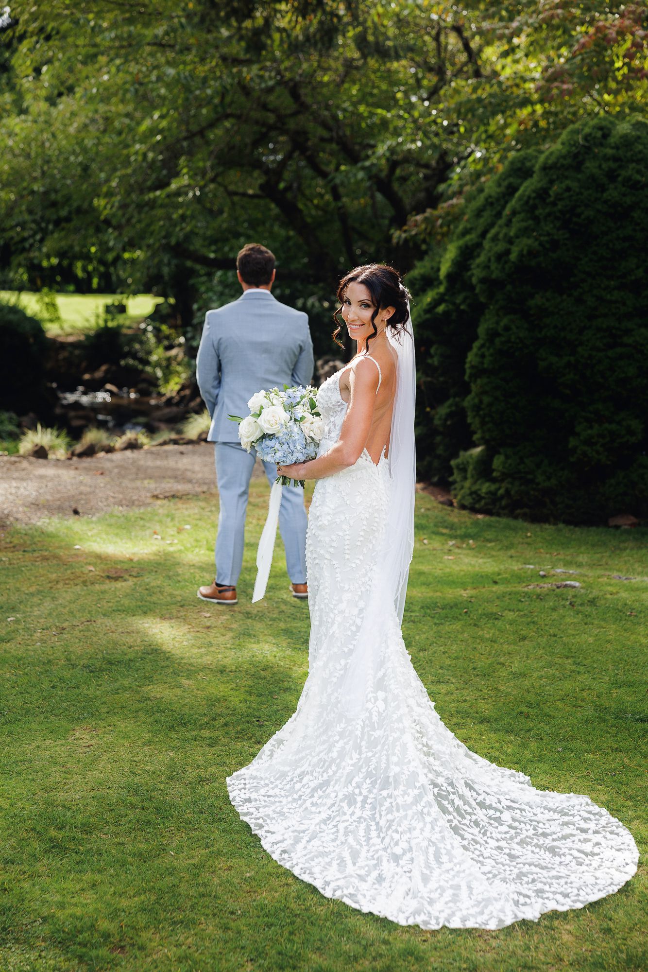 A wedding couple poses in a garden setting with the bride's lace dress train elegantly spread on the grass.