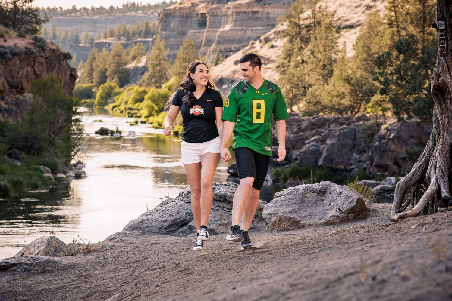 A couple walks together along a scenic river trail with rocky cliffs in the background.