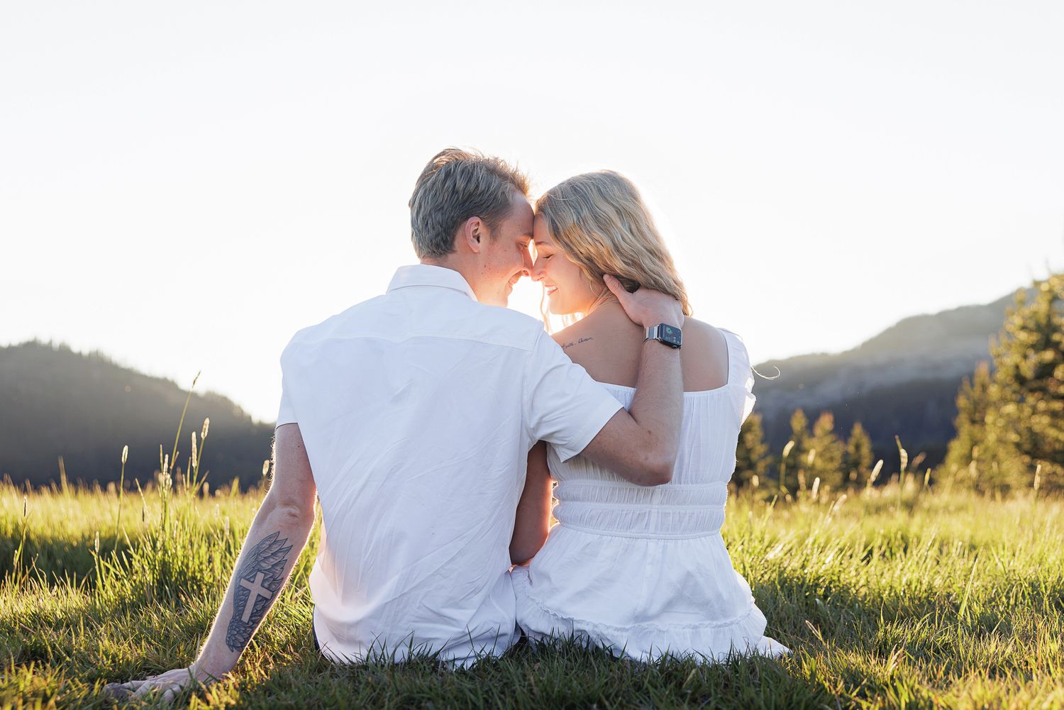 Couple sitting in a sunny meadow with mountain views, touching foreheads romantically in matching white outfits.