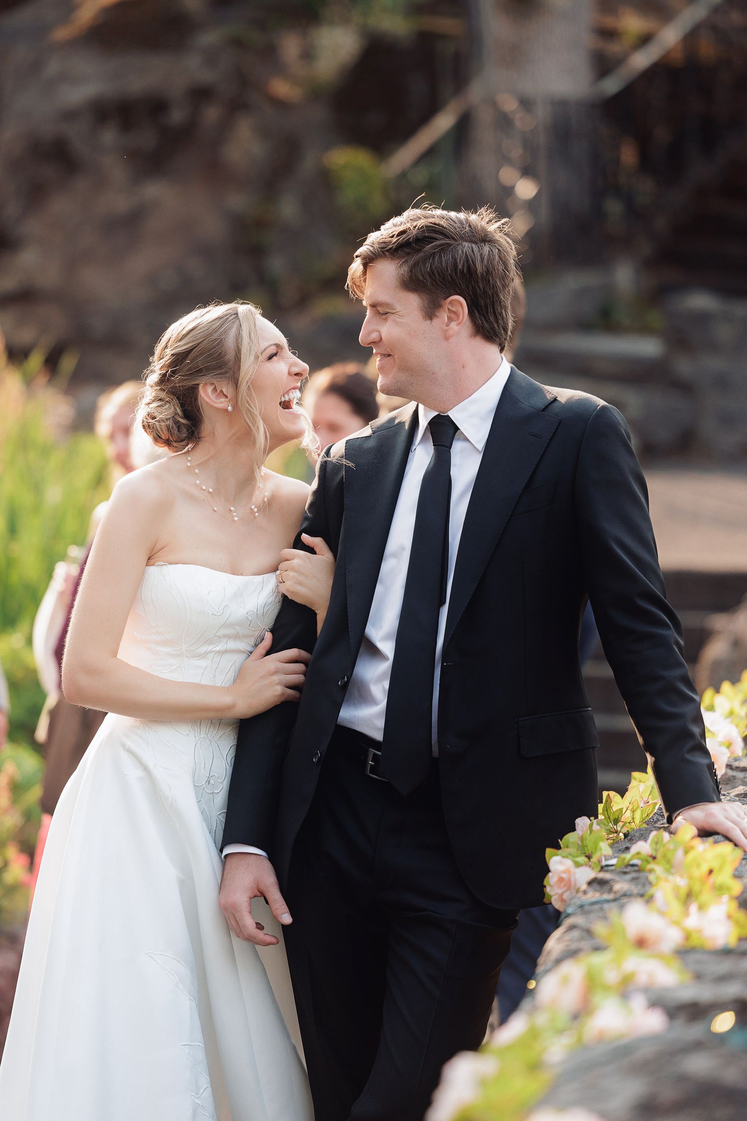 Bride and groom laughing together outdoors on their wedding day, she in a strapless gown, he in a black suit.