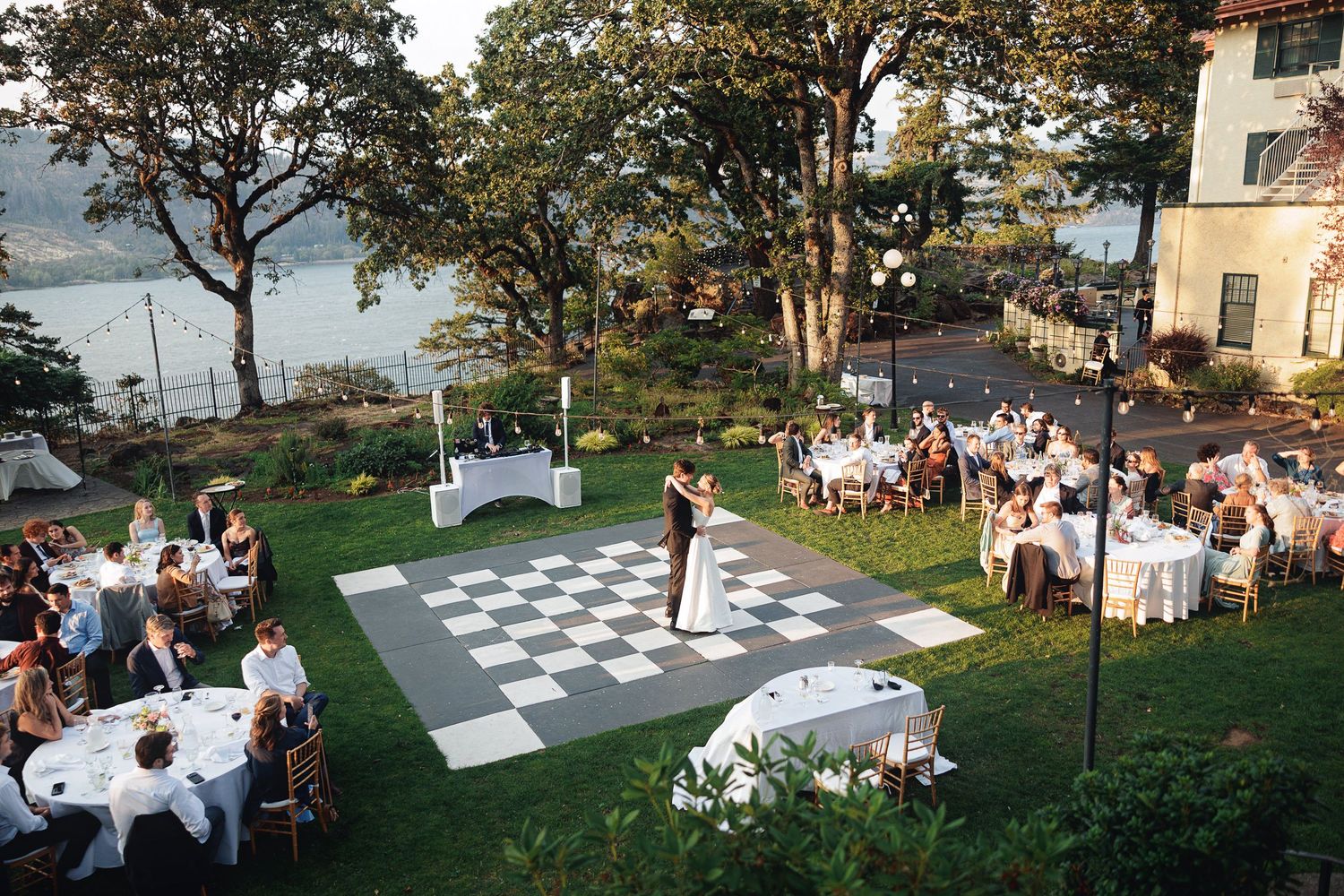 Outdoor wedding reception on checkered dance floor with couple dancing, guests at white tables, waterfront garden venue.