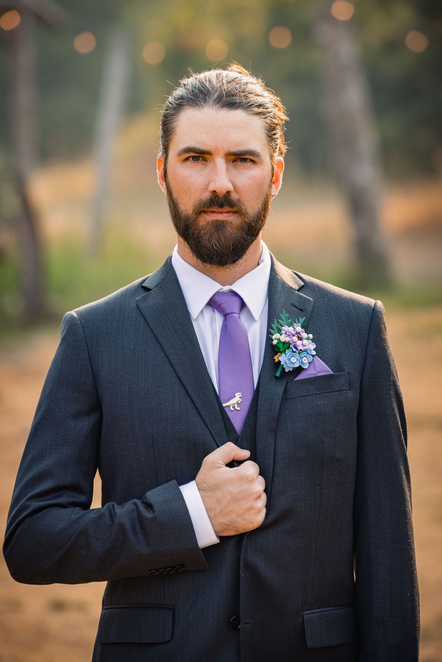 Groom in dark suit with purple tie and boutonniere poses outdoors with blurred bokeh background.