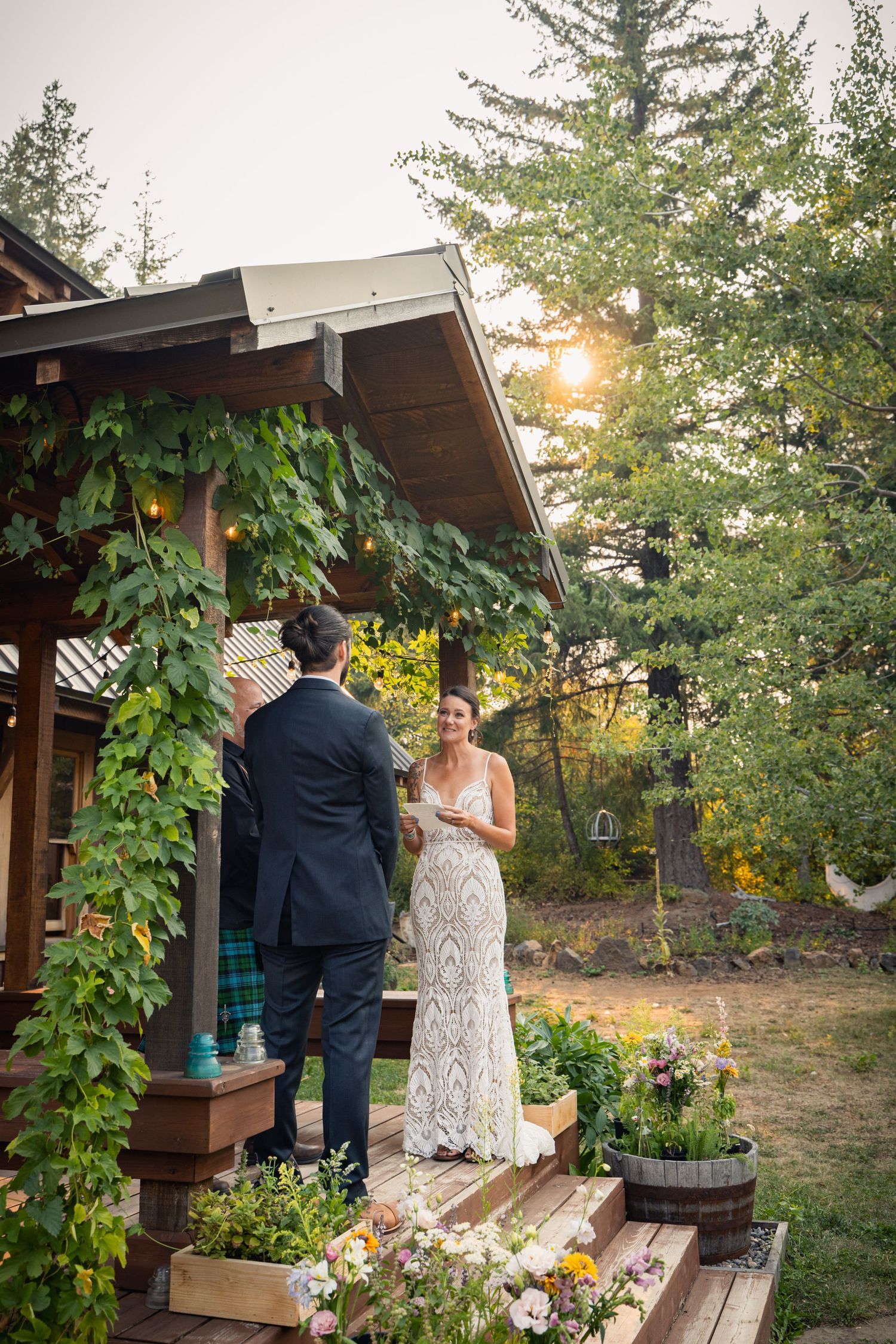 Two people stand on a rustic wooden deck adorned with climbing vines as golden sunlight filters through trees at sunset.