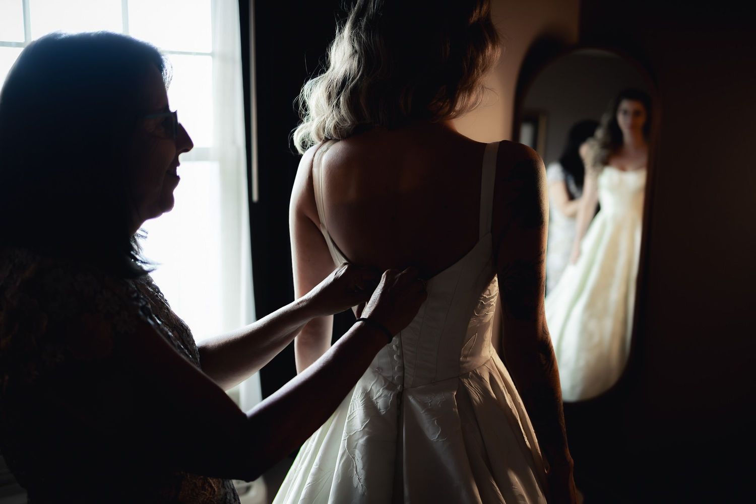 A silhouetted figure helps adjust a white wedding dress in atmospheric window lighting.
