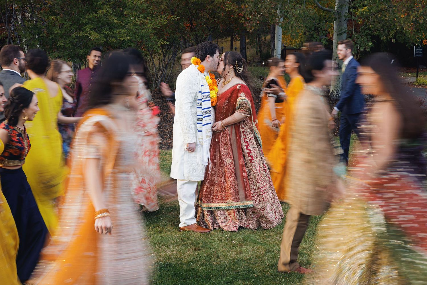 Indian bride and groom share a romantic moment surrounded by colorful guests during an outdoor wedding celebration.