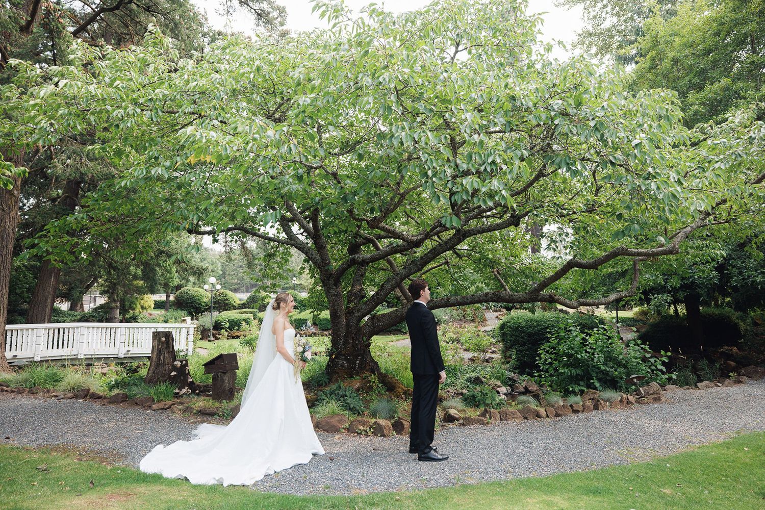 Bride and groom share a first look moment on a gravel path beneath a sprawling green tree at an elegant garden wedding venue.