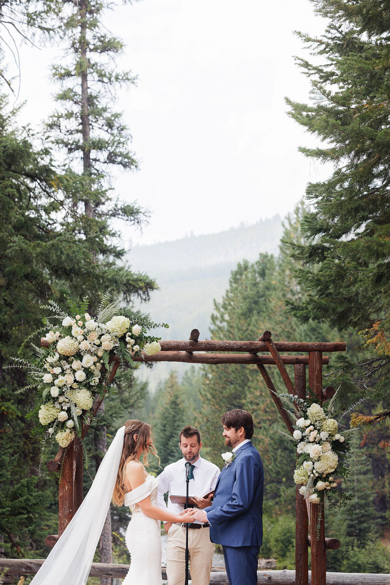 A wedding ceremony takes place under a rustic wooden arch decorated with white flowers in a mountain forest setting.
