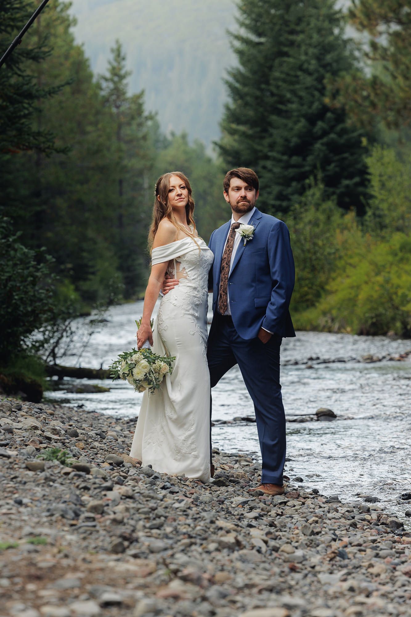A couple in wedding attire poses along a rocky riverbank surrounded by evergreen trees in a mountain setting.