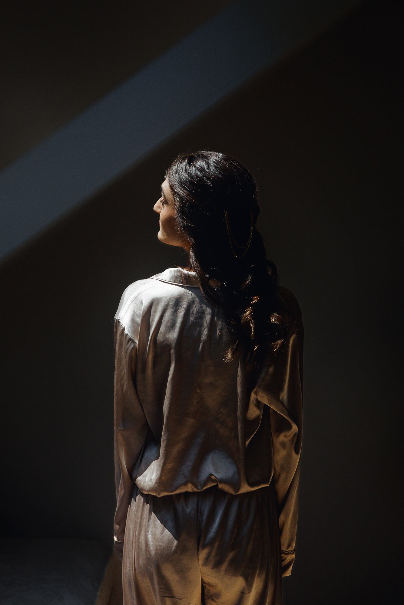 Woman in beige satin dress standing with back to camera in dramatic studio lighting with dark background.