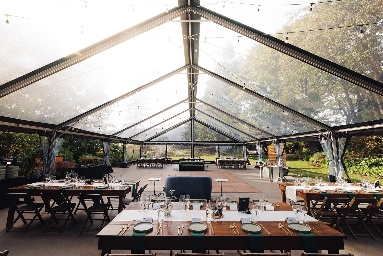 Elegant outdoor dining under a clear tent with wooden tables set for a formal event surrounded by lush greenery.