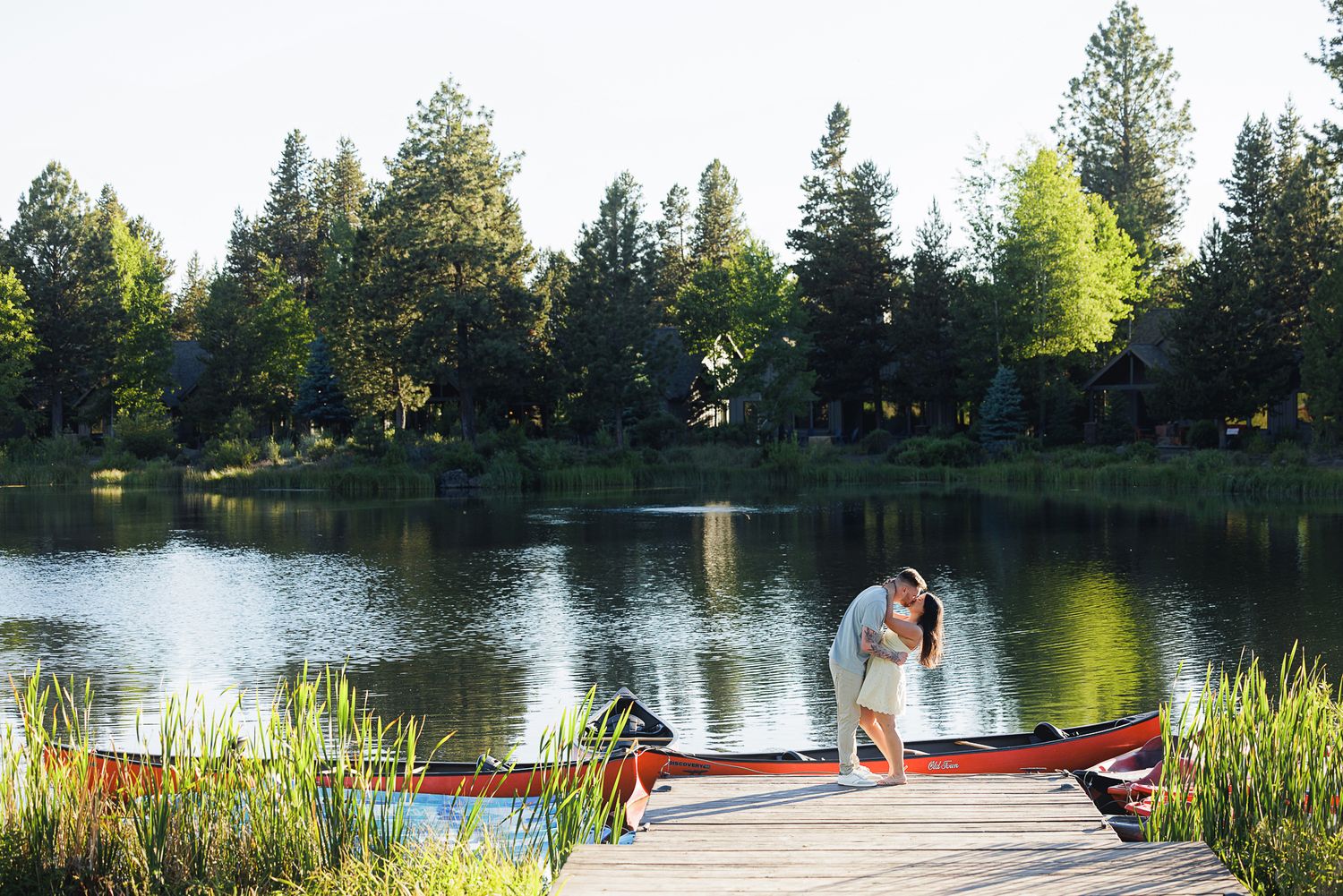 Couple sharing a romantic kiss on a wooden dock beside a red canoe on a serene lake surrounded by lush green trees.