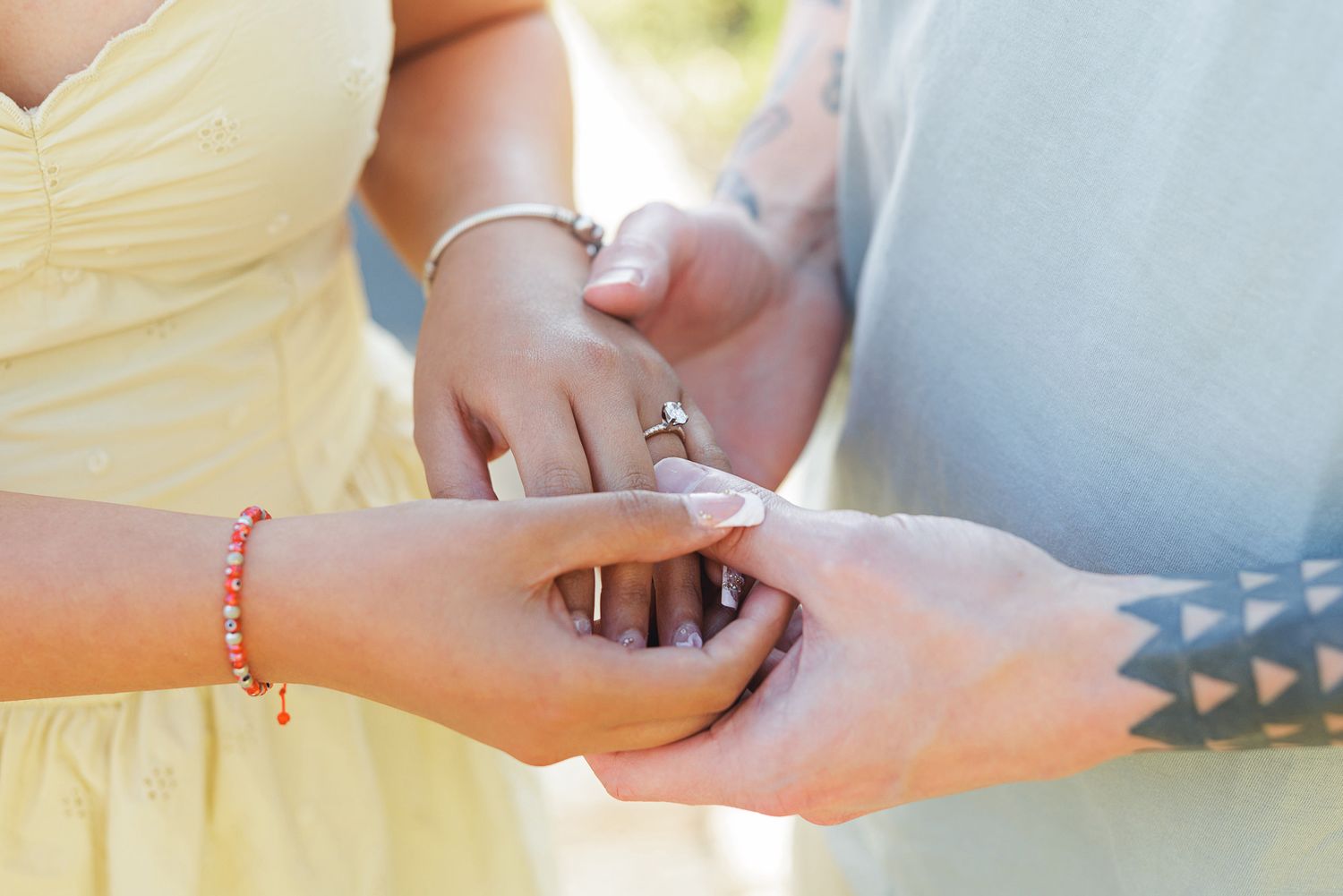 Couple holding hands during engagement ring exchange, woman wearing yellow dress and red bracelet, man with tattoos.