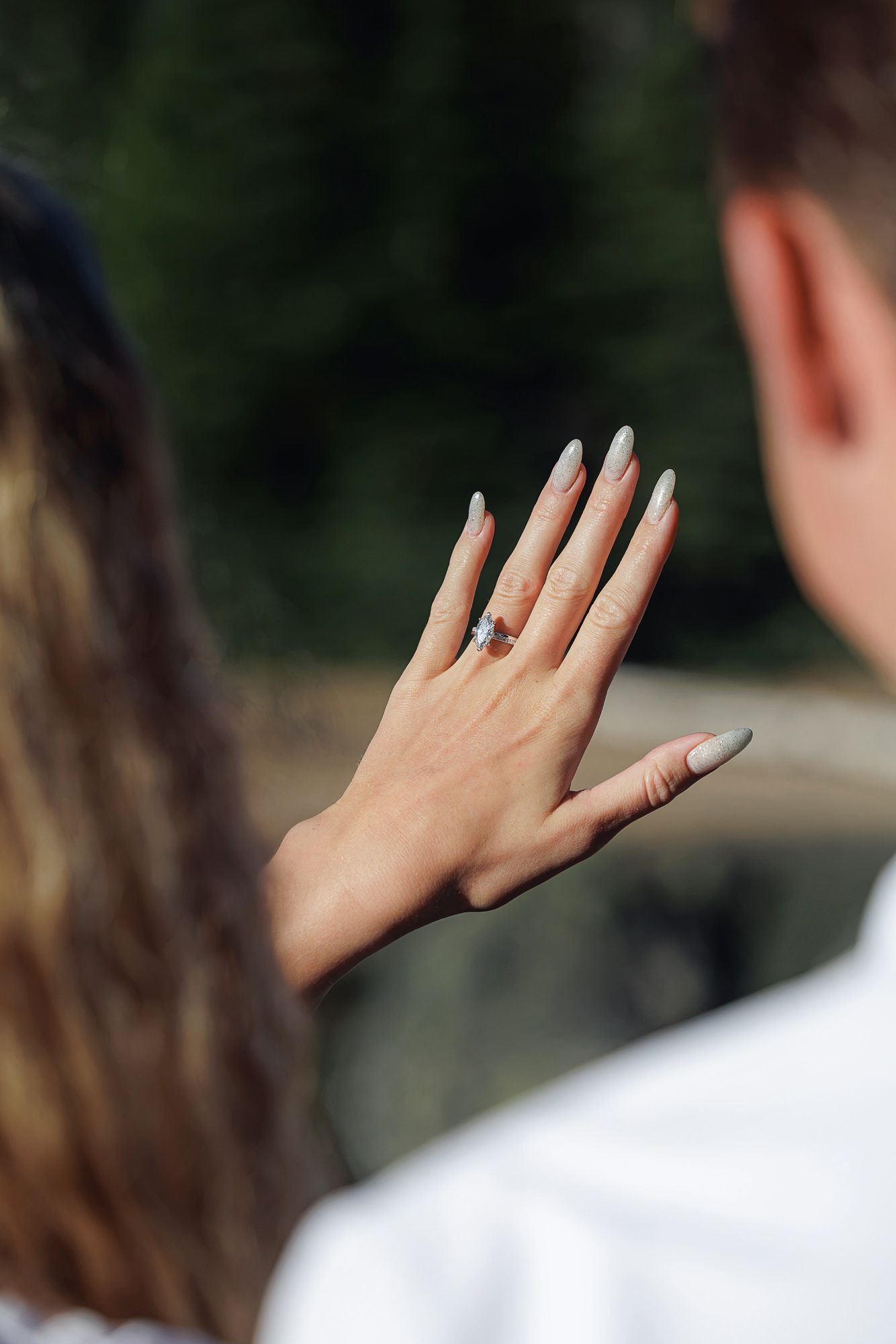 Woman displaying elegant engagement ring with blue gemstone on her hand while standing with her partner outdoors.