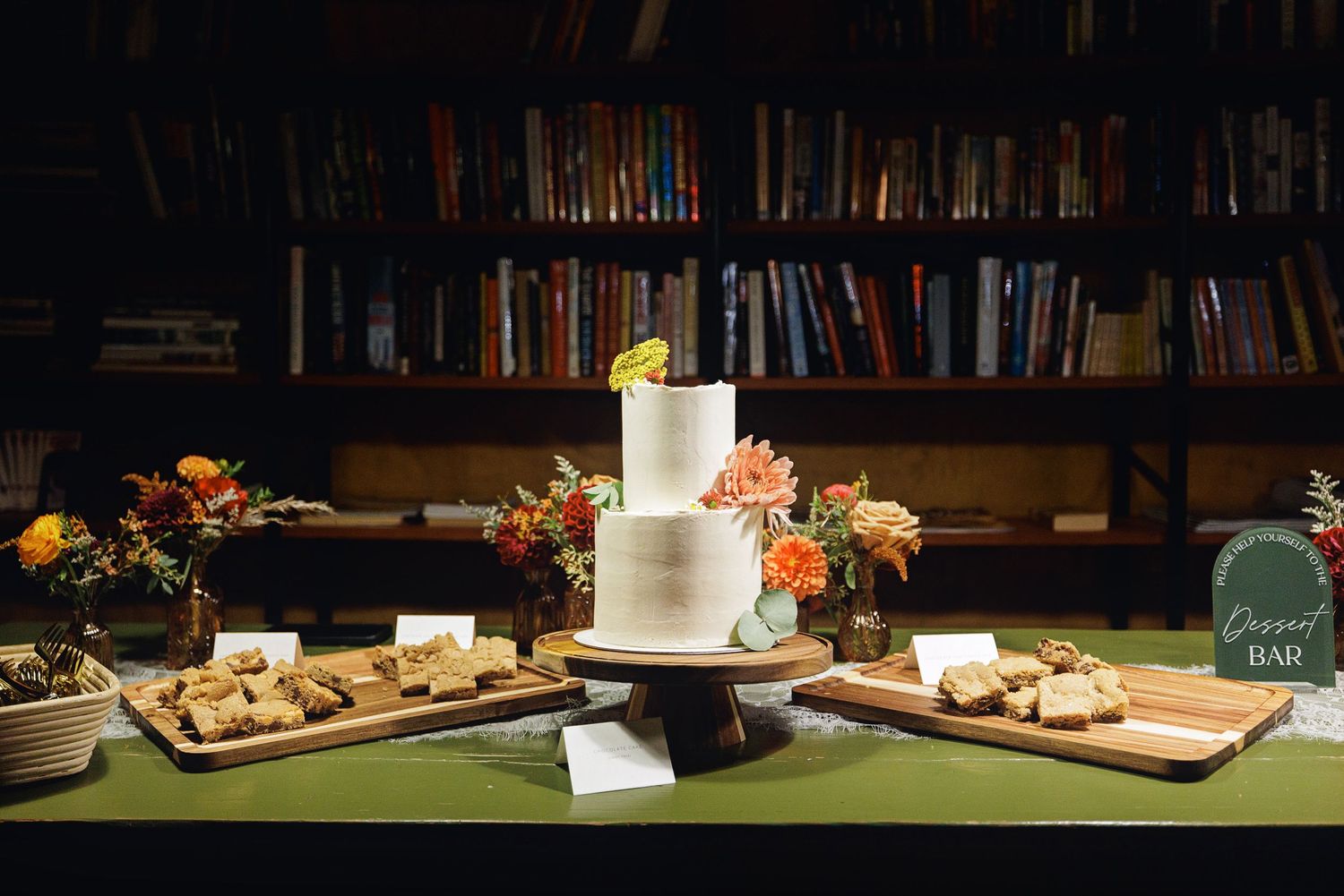 Elegant two-tier white wedding cake on a dessert bar table with floral arrangements and bookshelves backdrop.