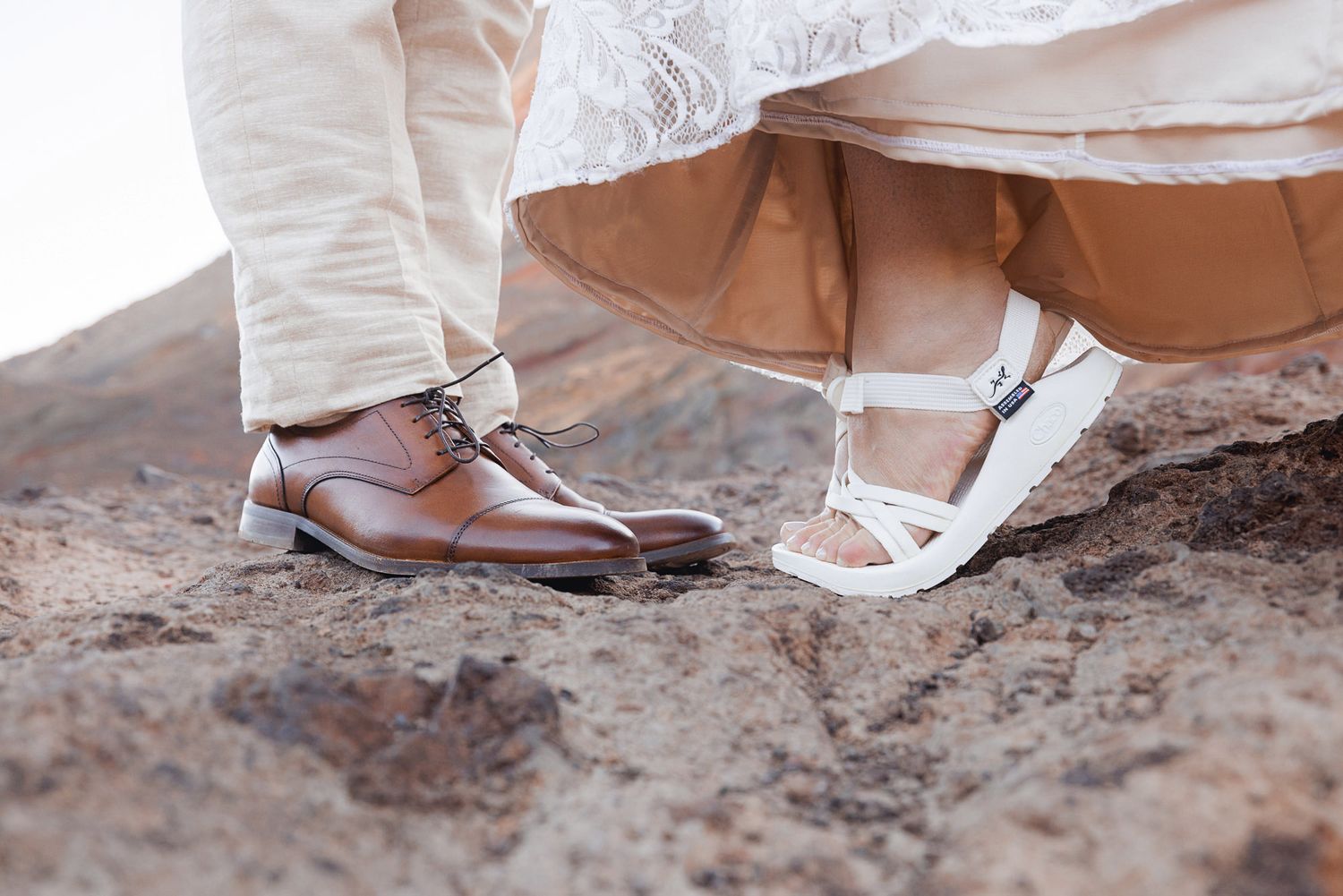 Close-up of a groom's brown leather dress shoes and a bride's white sandals standing on rugged rocky terrain at an outdoor wedding.
