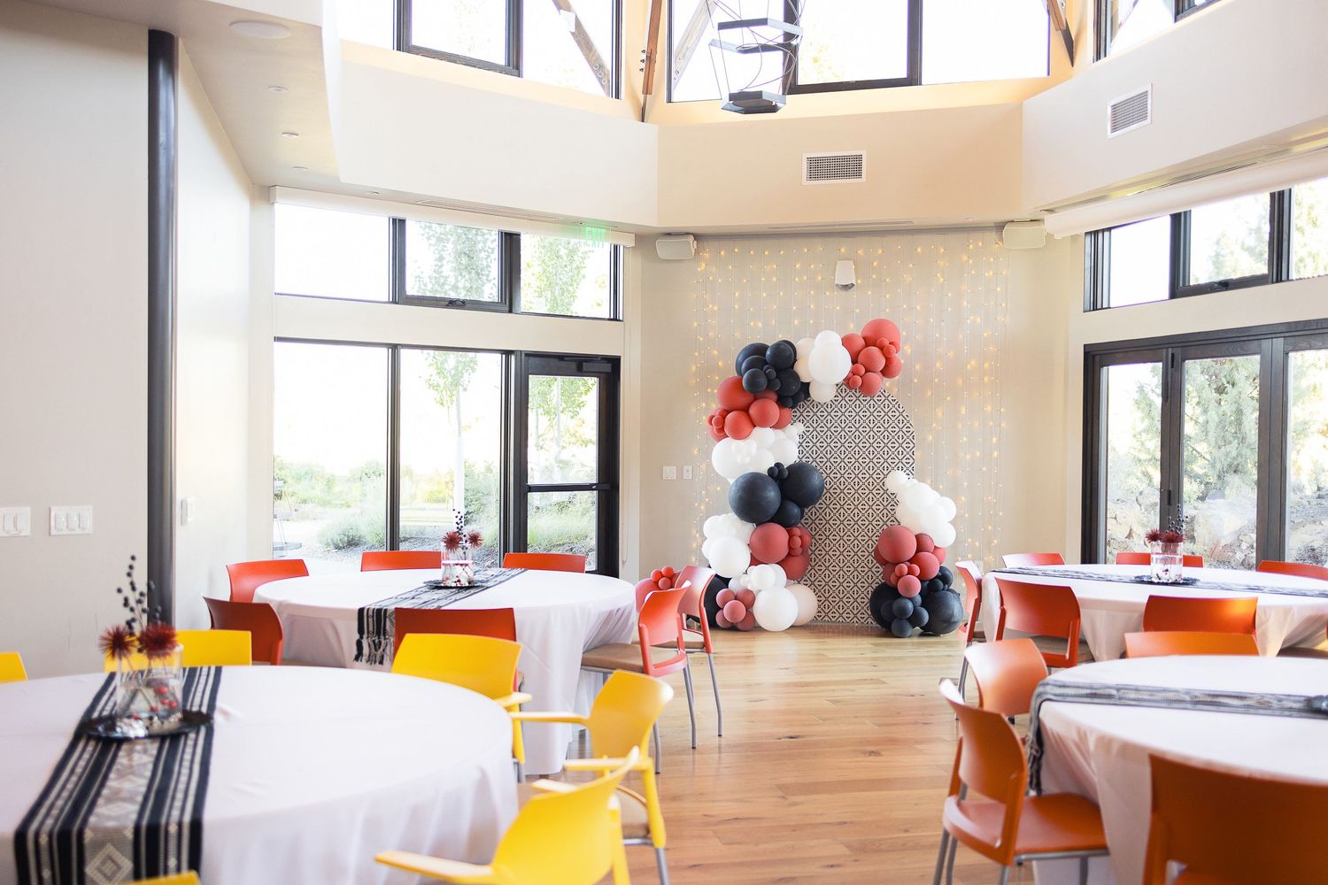 Modern event space with white tables, yellow chairs and red balloon decorations near tall windows.