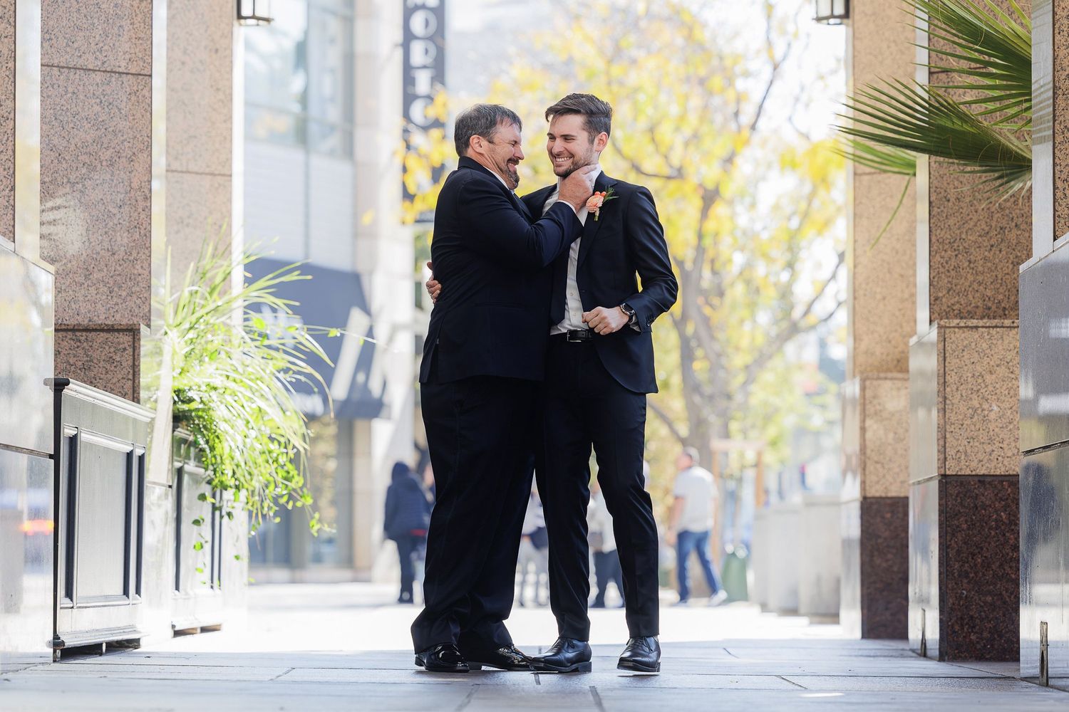 Two people in black formal attire share a happy moment on an elegant outdoor street with columns and trees.
