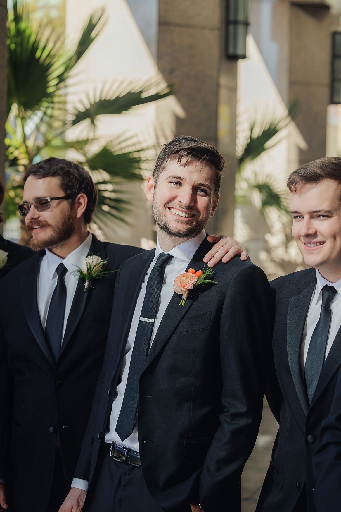 Three groomsmen in black suits and ties stand together smiling outdoors with palm trees visible in the background.