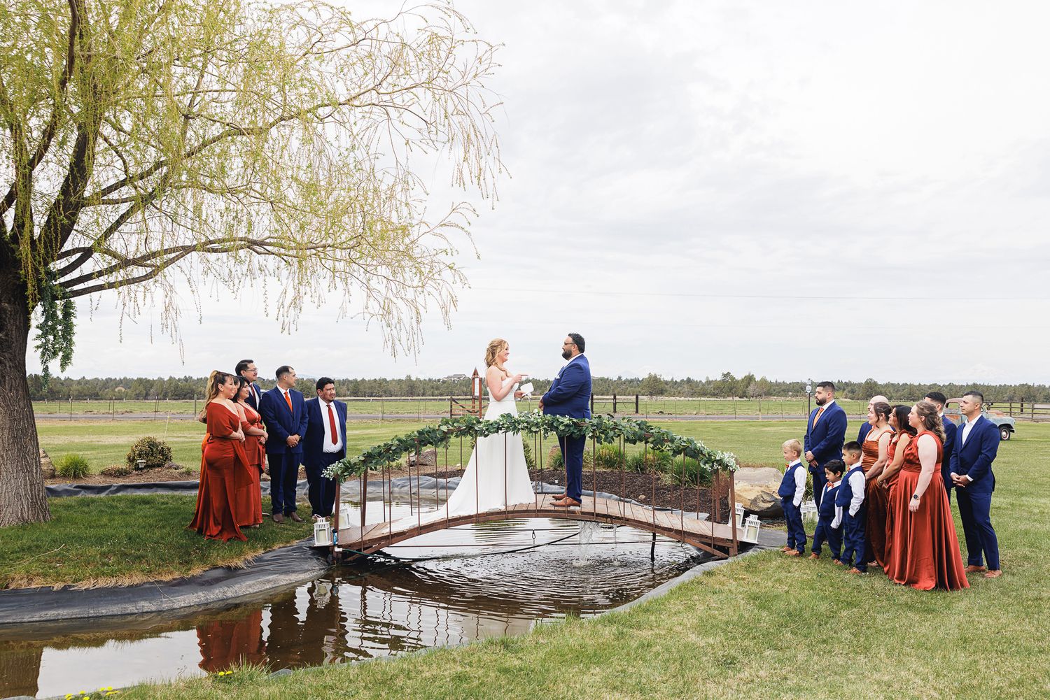 Wedding ceremony on a bridge over a pond with bridal party in red and green dresses at an outdoor venue.