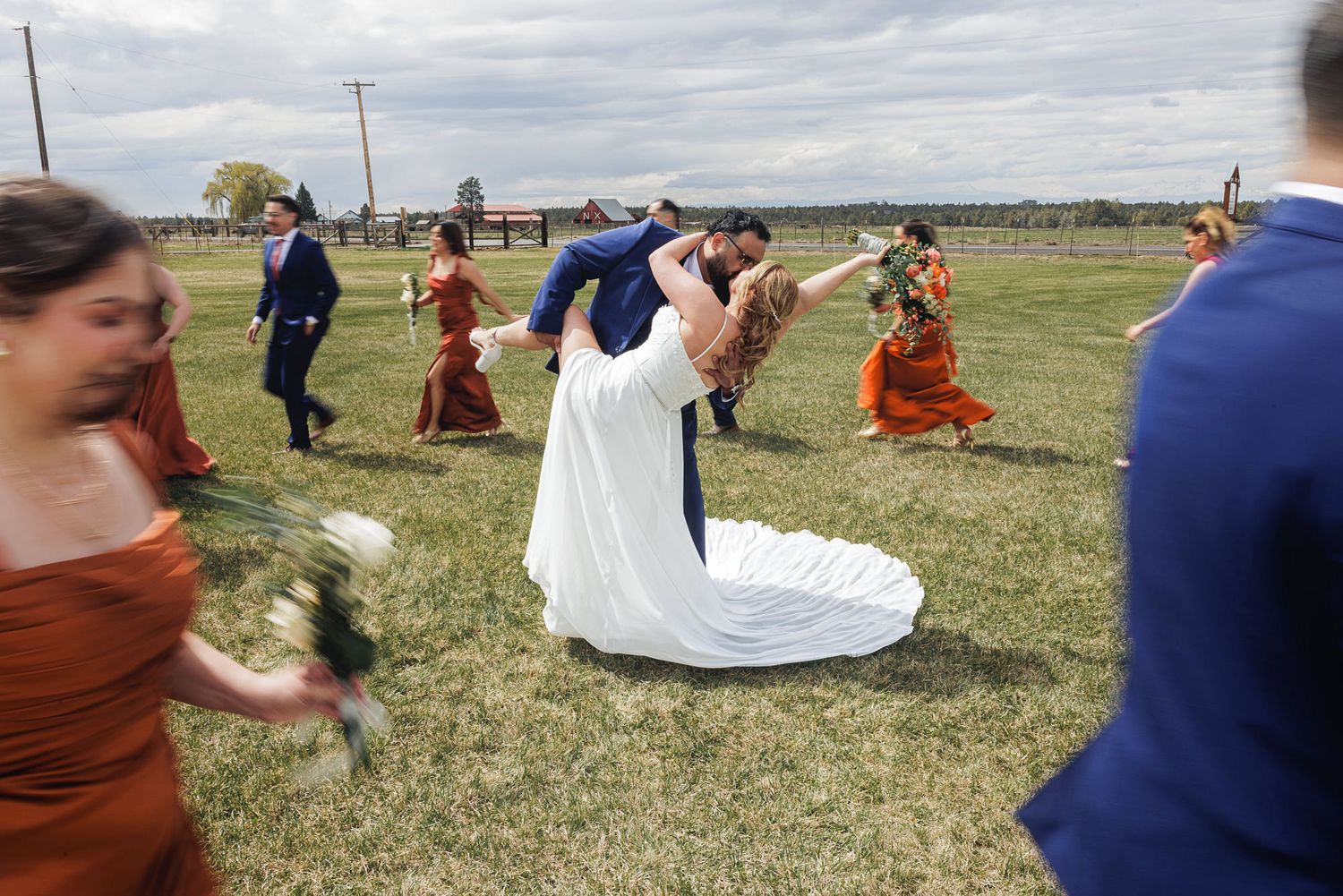 Bride and groom share a romantic dip kiss surrounded by wedding party in rust and blue attire on green lawn.