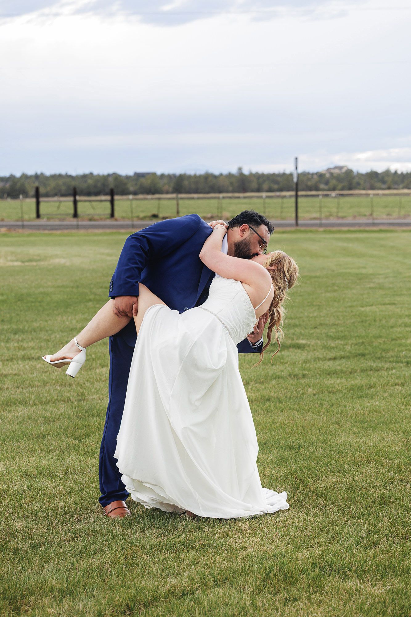 Groom in navy suit dips and kisses bride in white gown on green grass field during outdoor wedding celebration.