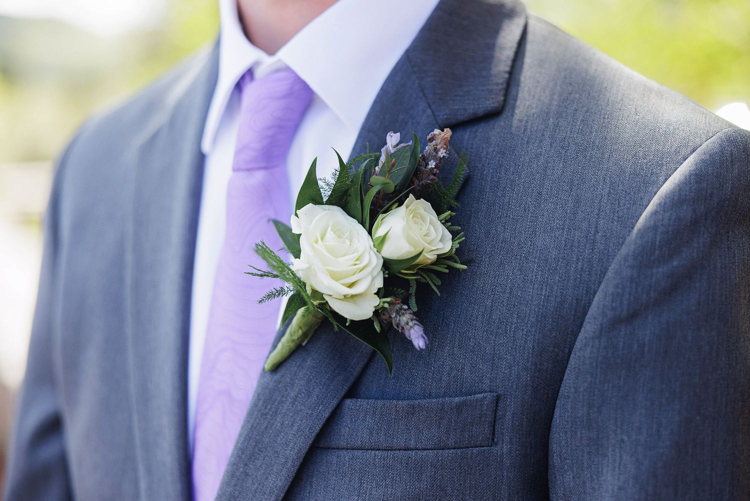 Groom wearing gray suit with lavender tie and white rose boutonniere with greenery on wedding day.