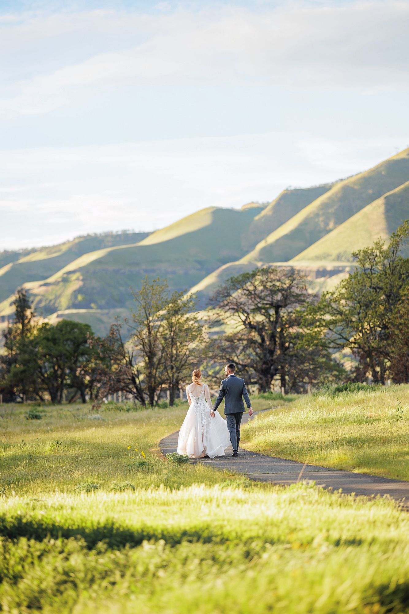 Bride and groom walking hand in hand on a path through lush green meadows with scenic rolling hills backdrop.