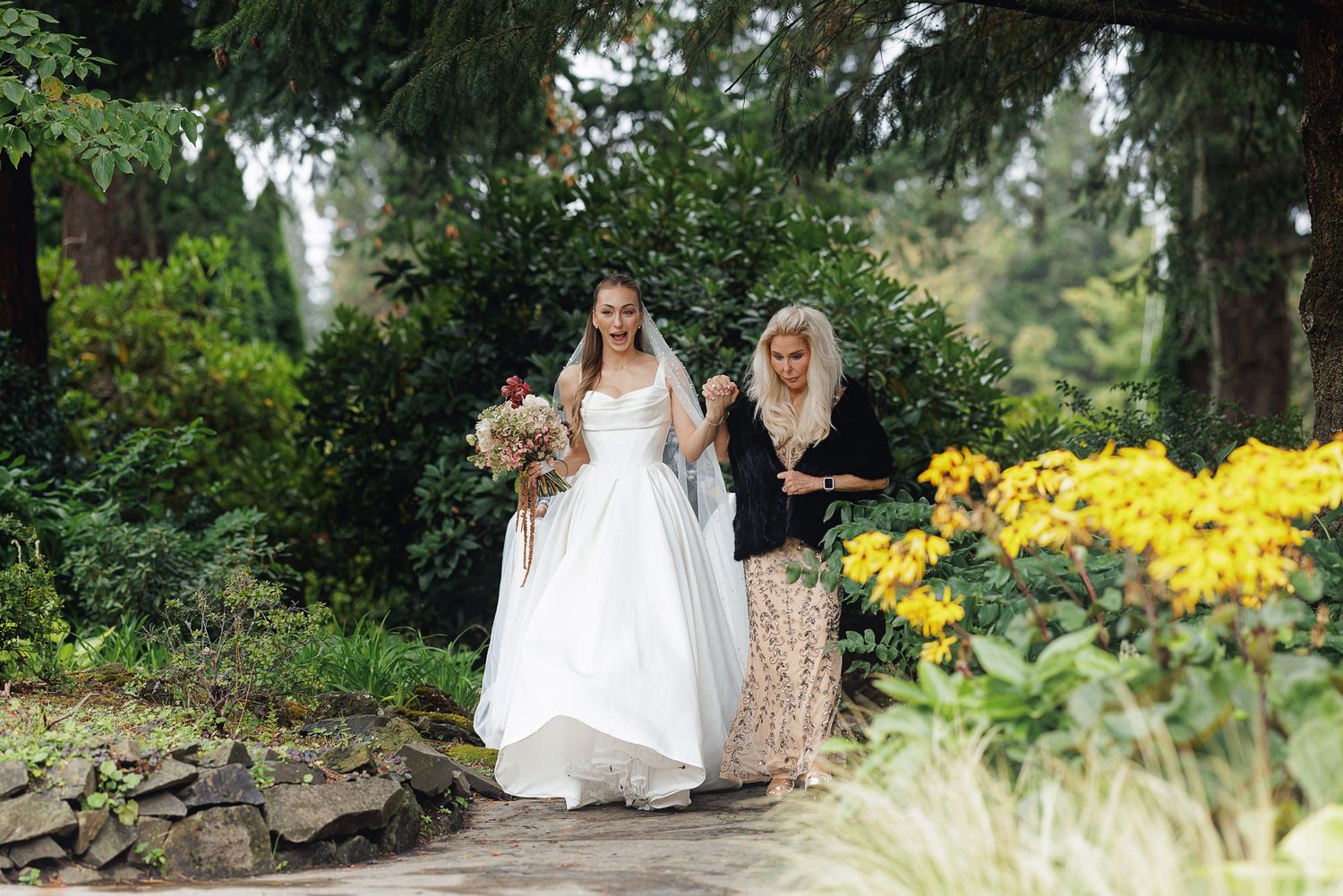 Bride in white gown holding bouquet walks with guest through lush garden path with yellow flowers.
