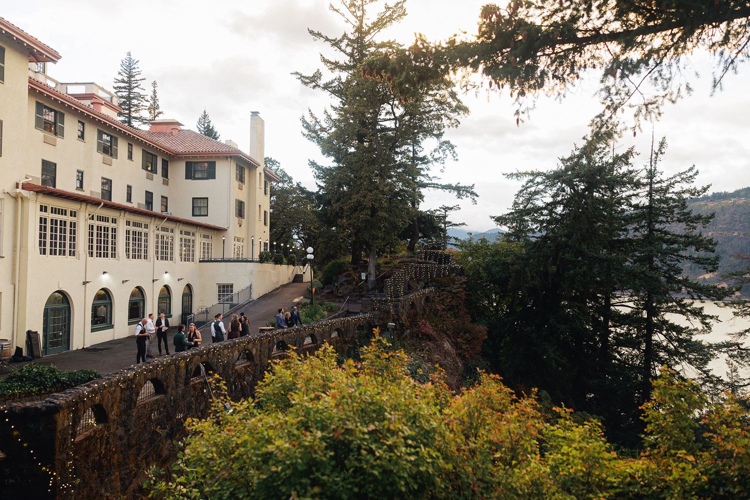 Historic yellow hotel perched on a cliff overlooking a river, surrounded by tall evergreen trees and autumn foliage.