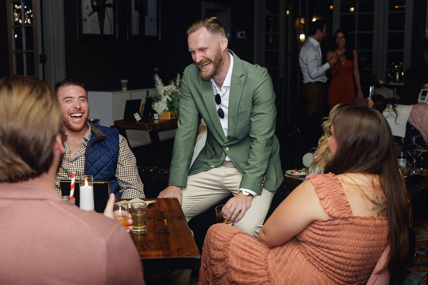 Man in green blazer laughing with friends at a restaurant dinner party in a dimly lit venue.
