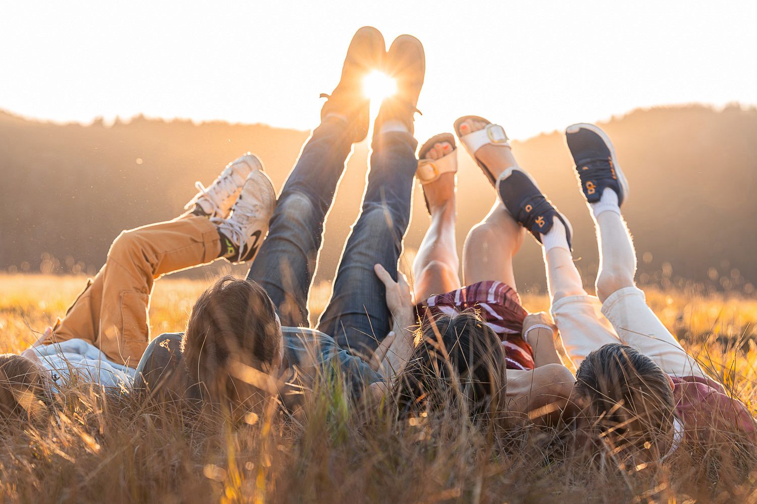 Friends lying in grass field raising legs together against golden sunset sky in sunny summer evening.