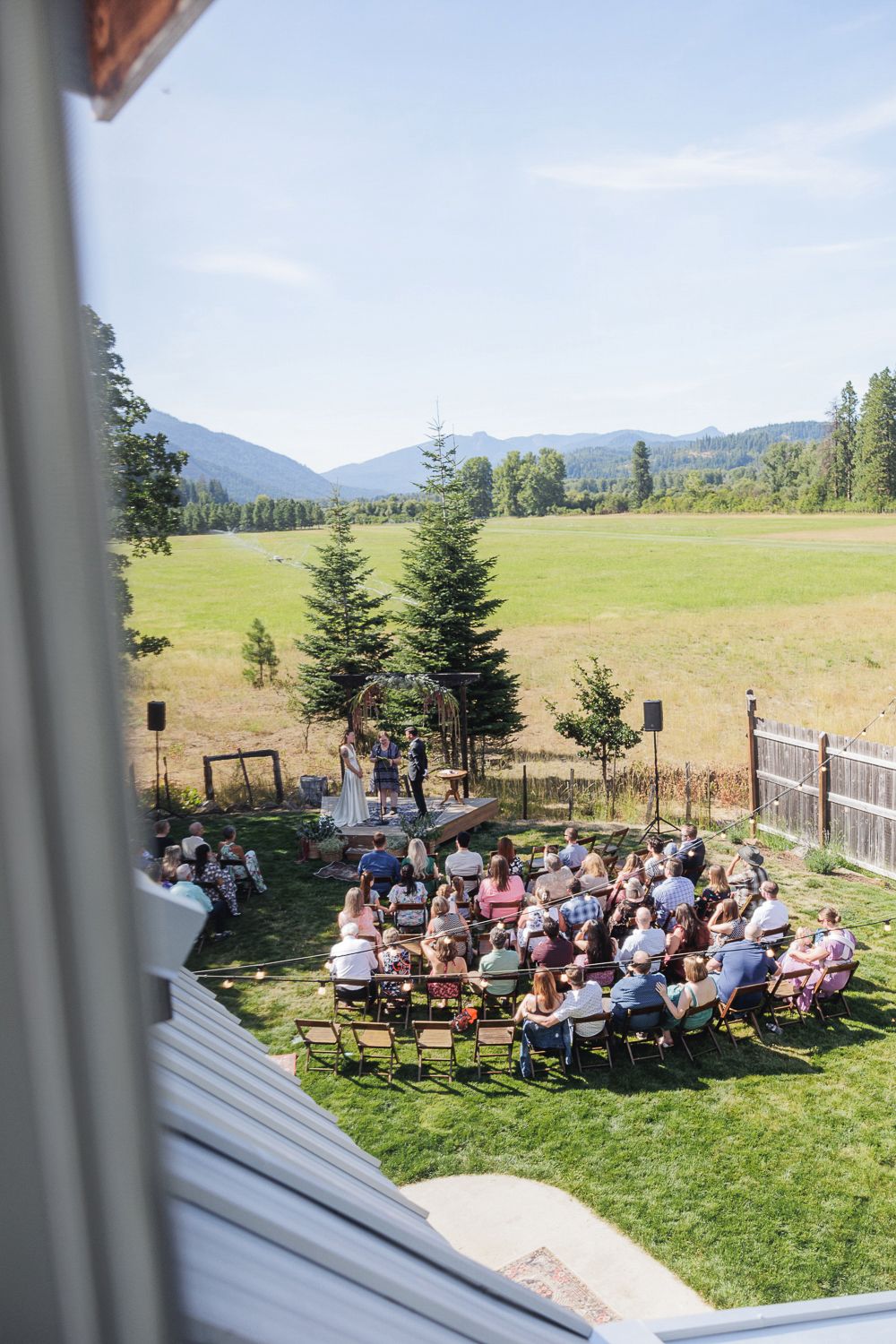 Outdoor wedding ceremony in a mountain meadow with guests seated in white chairs facing an evergreen backdrop.