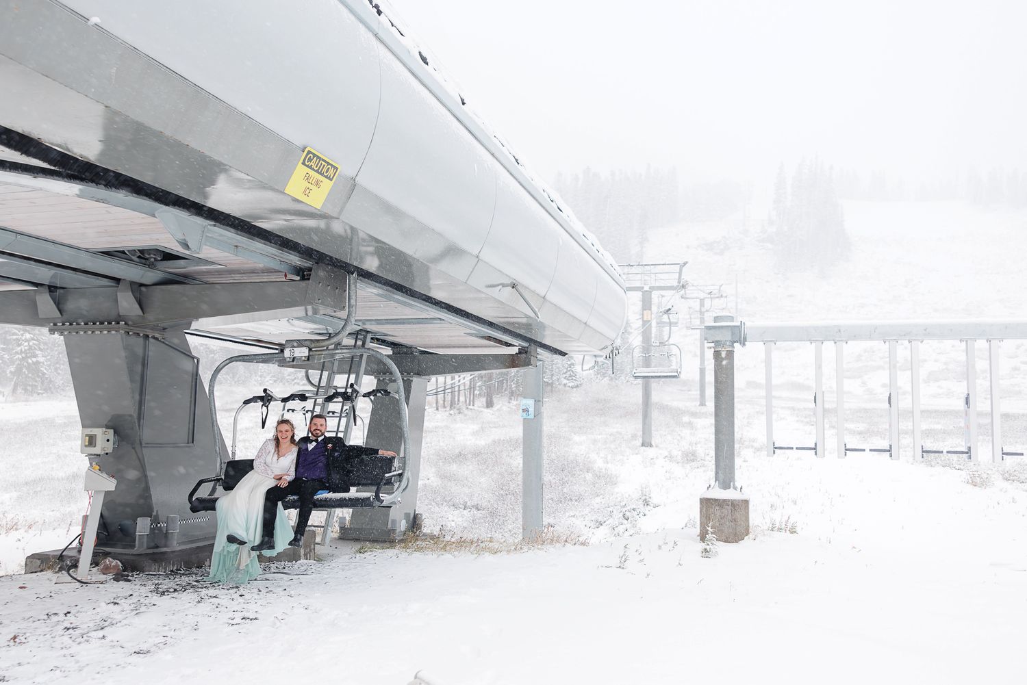 Skiers ride a chairlift through heavy snowfall at a snowy mountain resort on a foggy winter day.