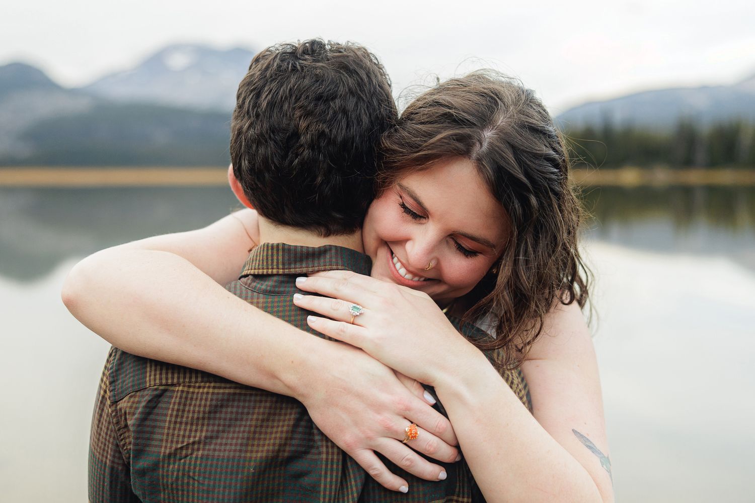 A couple shares an intimate embrace while standing near a scenic lake with mountains in the background.