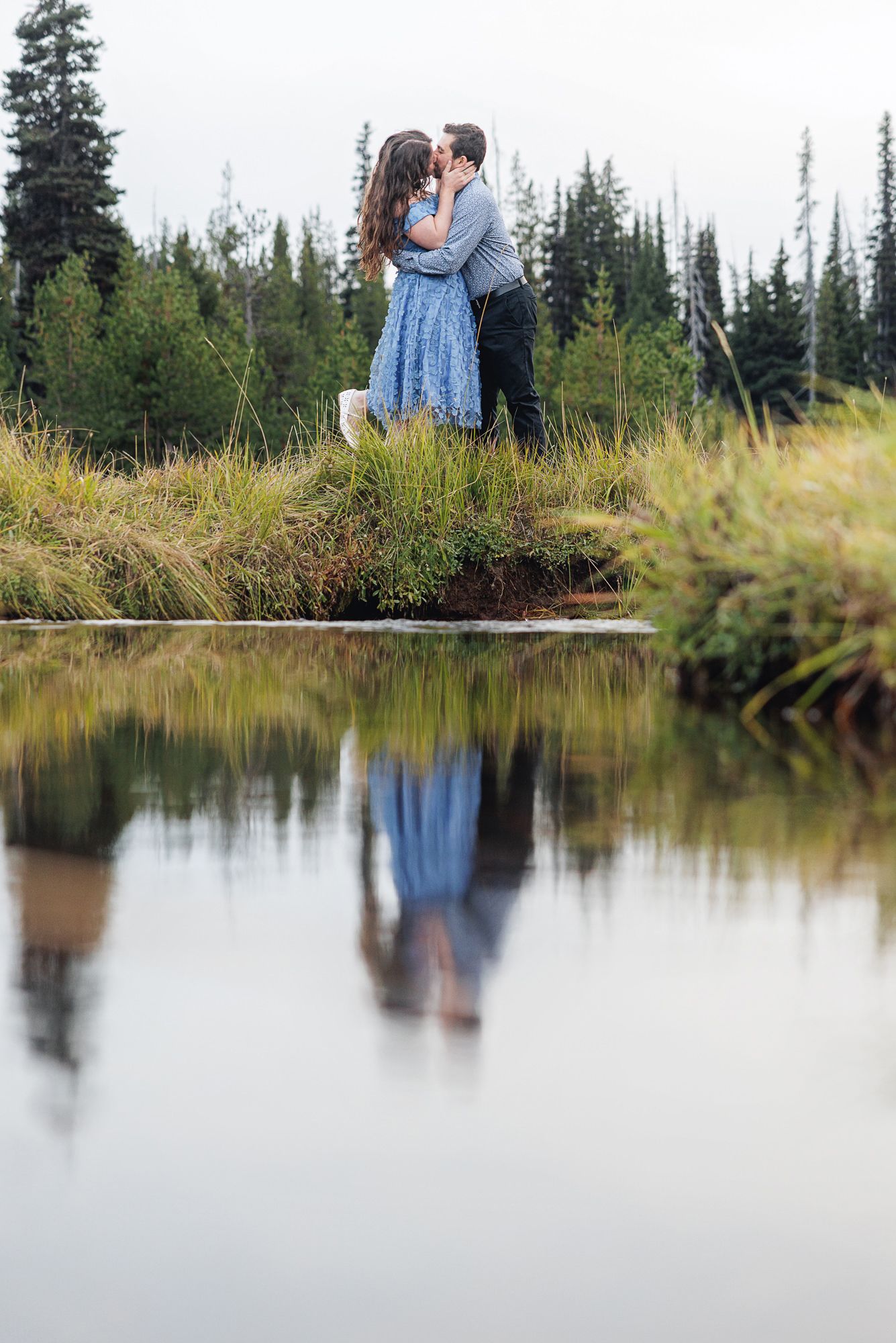 A romantic couple embraces on a grassy hill overlooking a still lake with evergreen trees in the background.