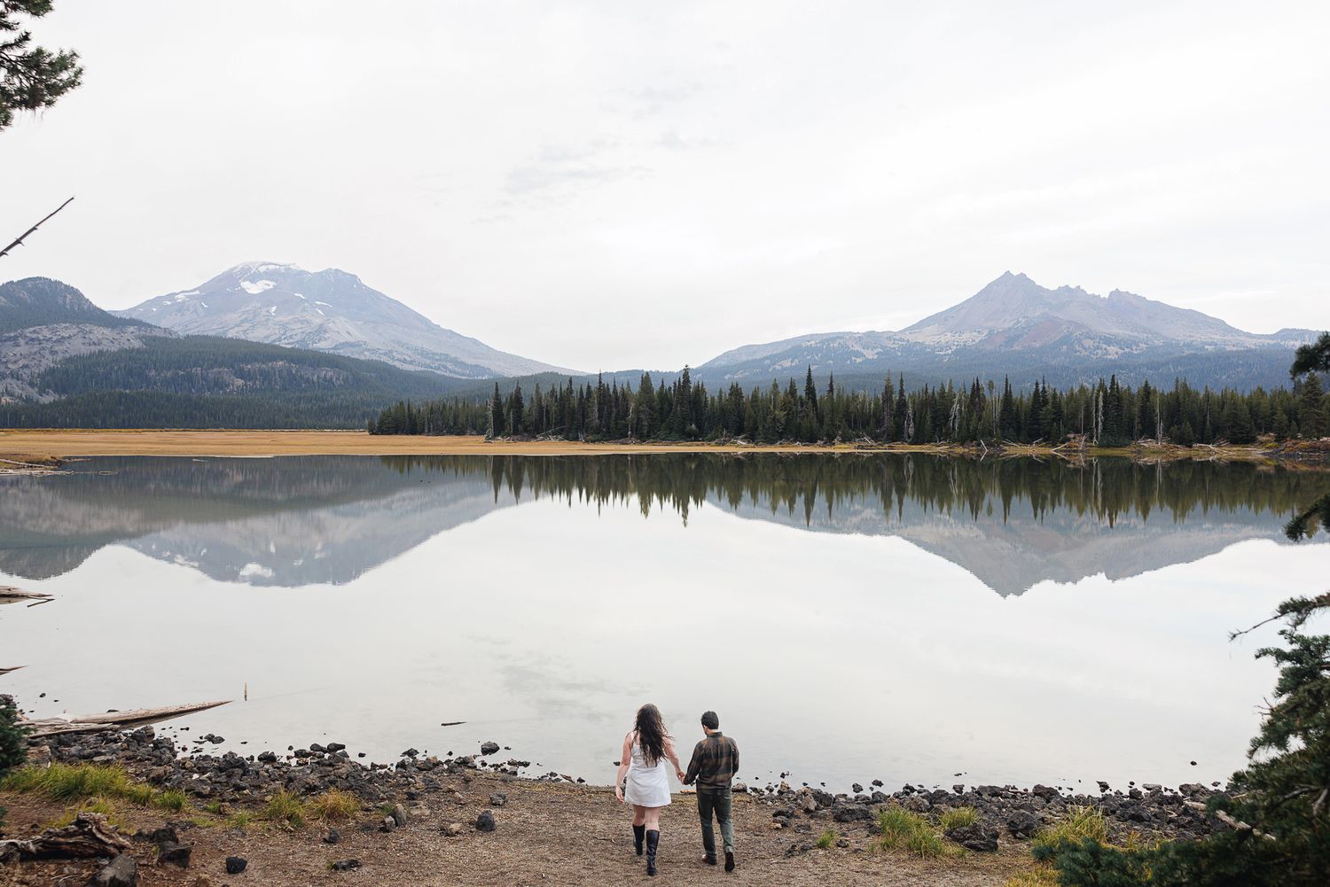 Scenic panoramic view of a serene mountain lake surrounded by evergreen forests and snow-capped peaks in the distance.