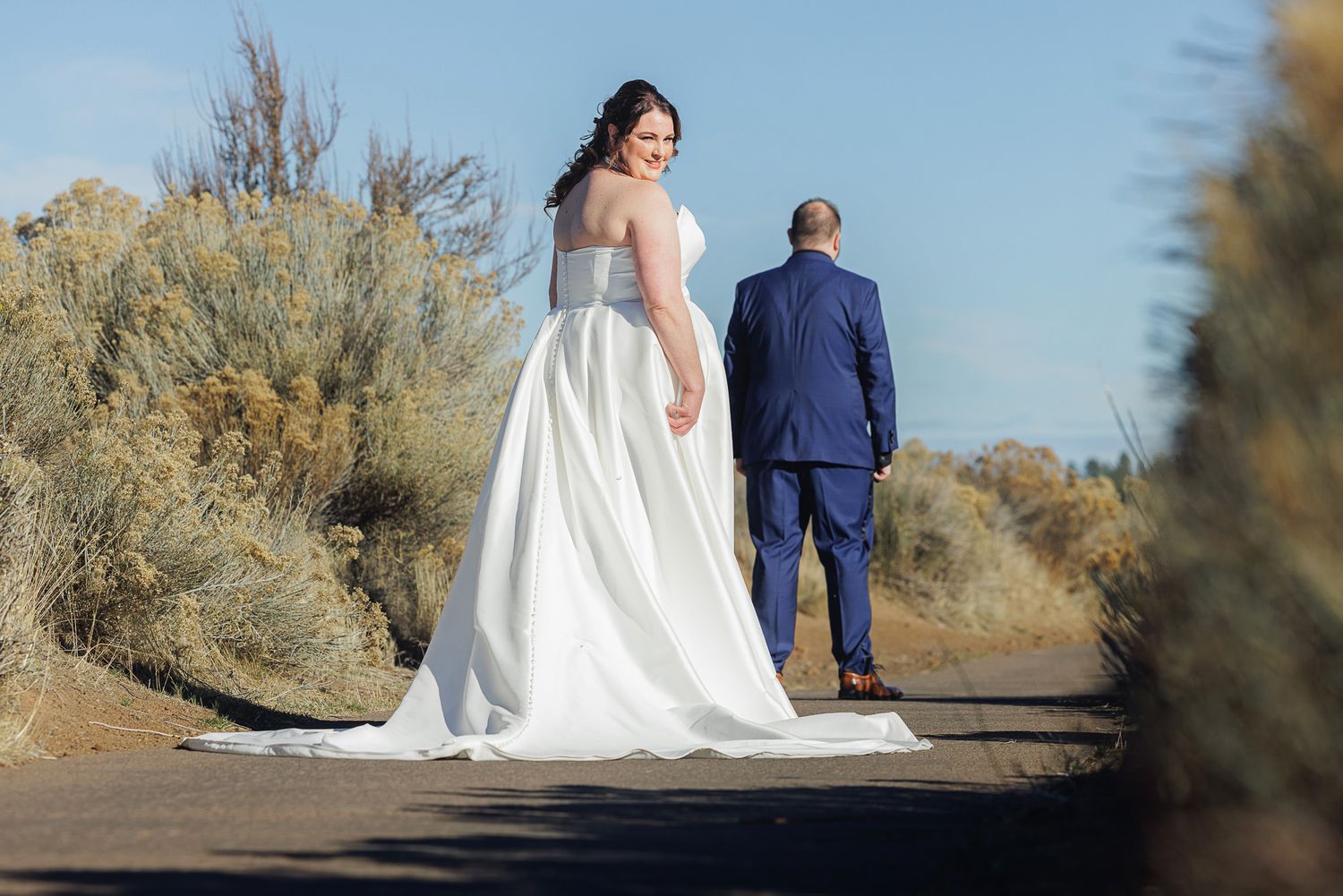 A couple in wedding attire walks along a desert path surrounded by desert brush and golden light.