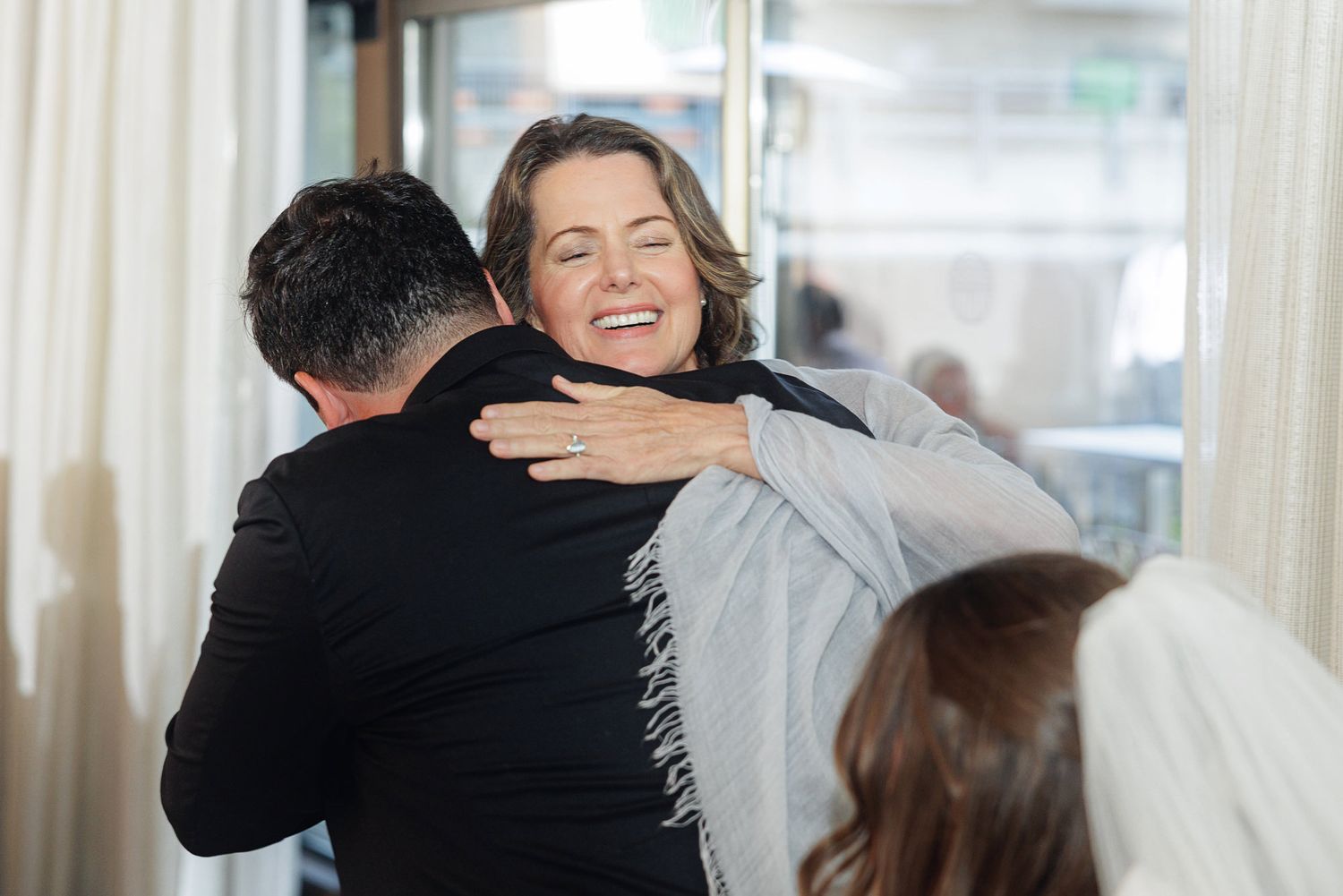 Two people sharing a warm hug at a wedding event, with a woman smiling joyfully wearing a gray scarf.