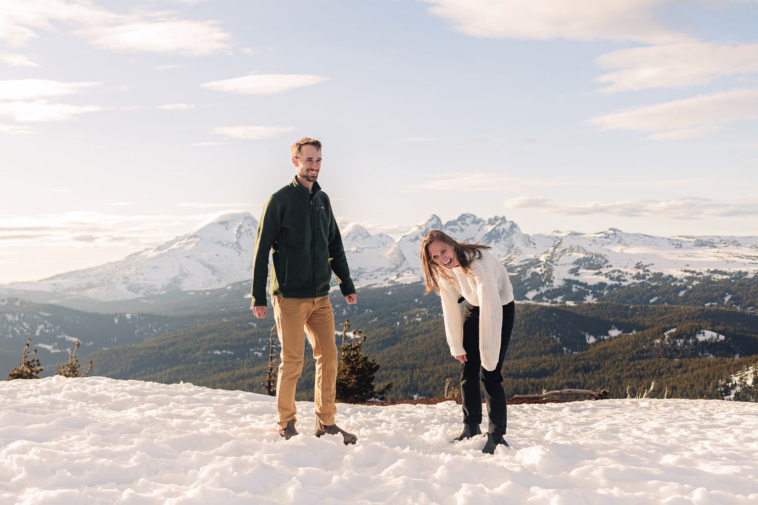 Couple posing on snowy mountain summit with scenic Rocky Mountain winter landscape and pine forests in background.