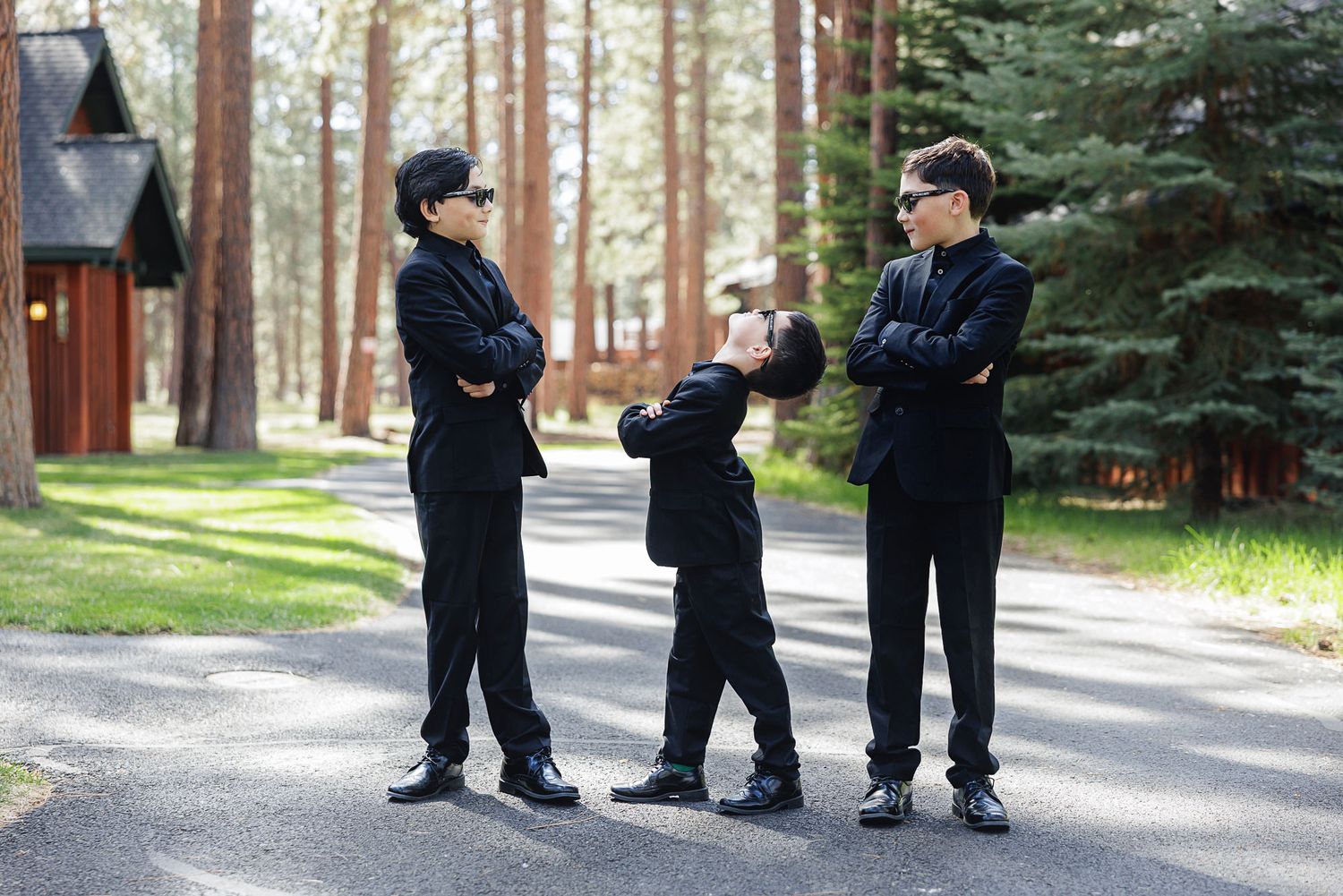 Three boys in matching black suits and sunglasses stand on a path in a wooded park, arms crossed confidently.
