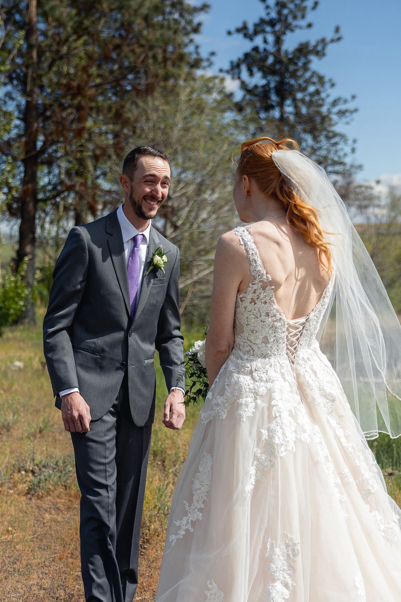 A smiling groom in a gray suit with purple tie faces his red-haired bride in a lace wedding dress outdoors.