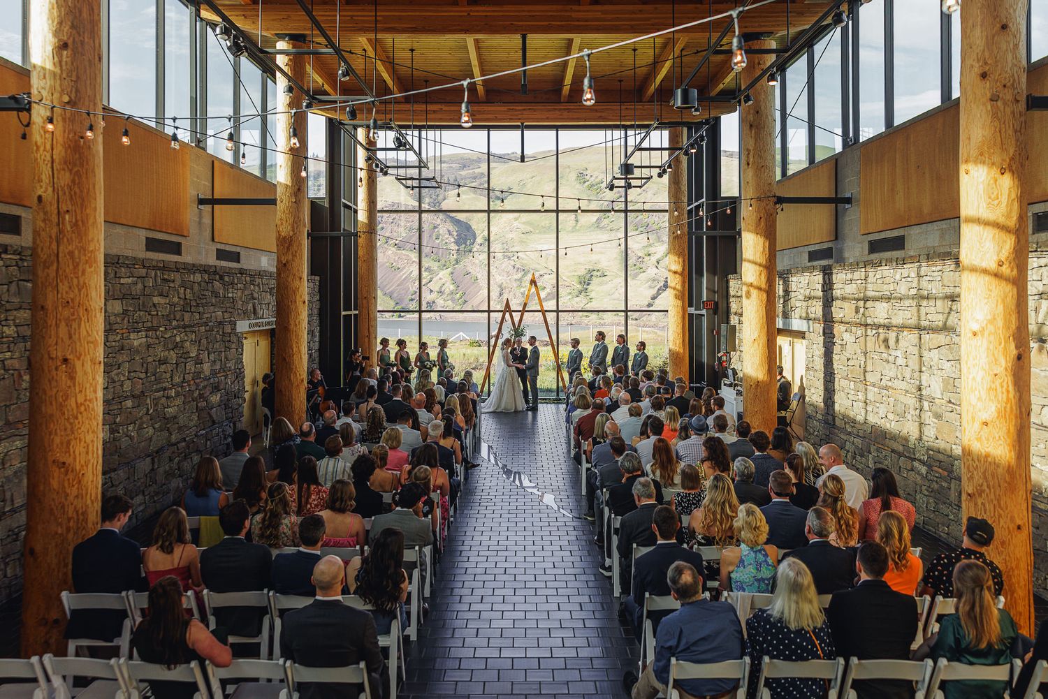 Indoor wedding ceremony in a rustic venue with large windows, stone walls, wooden beams, and mountain views.