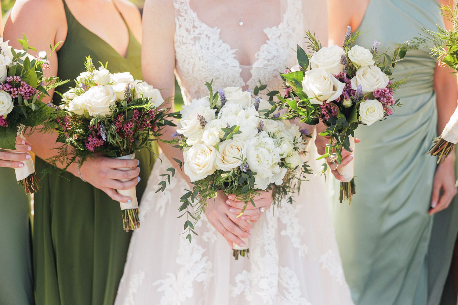 Bride holding white rose bouquet flanked by bridesmaids in sage green dresses with purple and white floral arrangements.