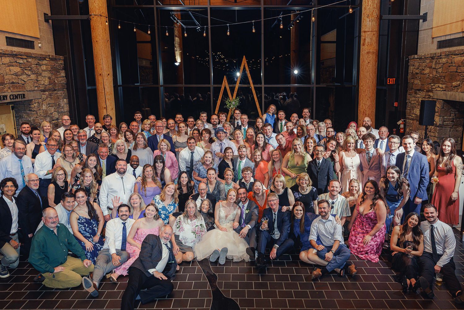 Large wedding group photo with guests posing together in elegant venue with stone walls and string lights.